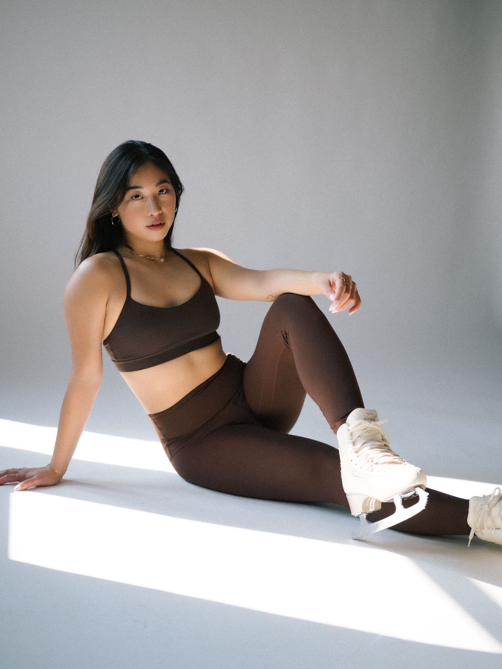Person in brown athletic wear and white sneakers posing on floor in minimalist setting with dramatic lighting.