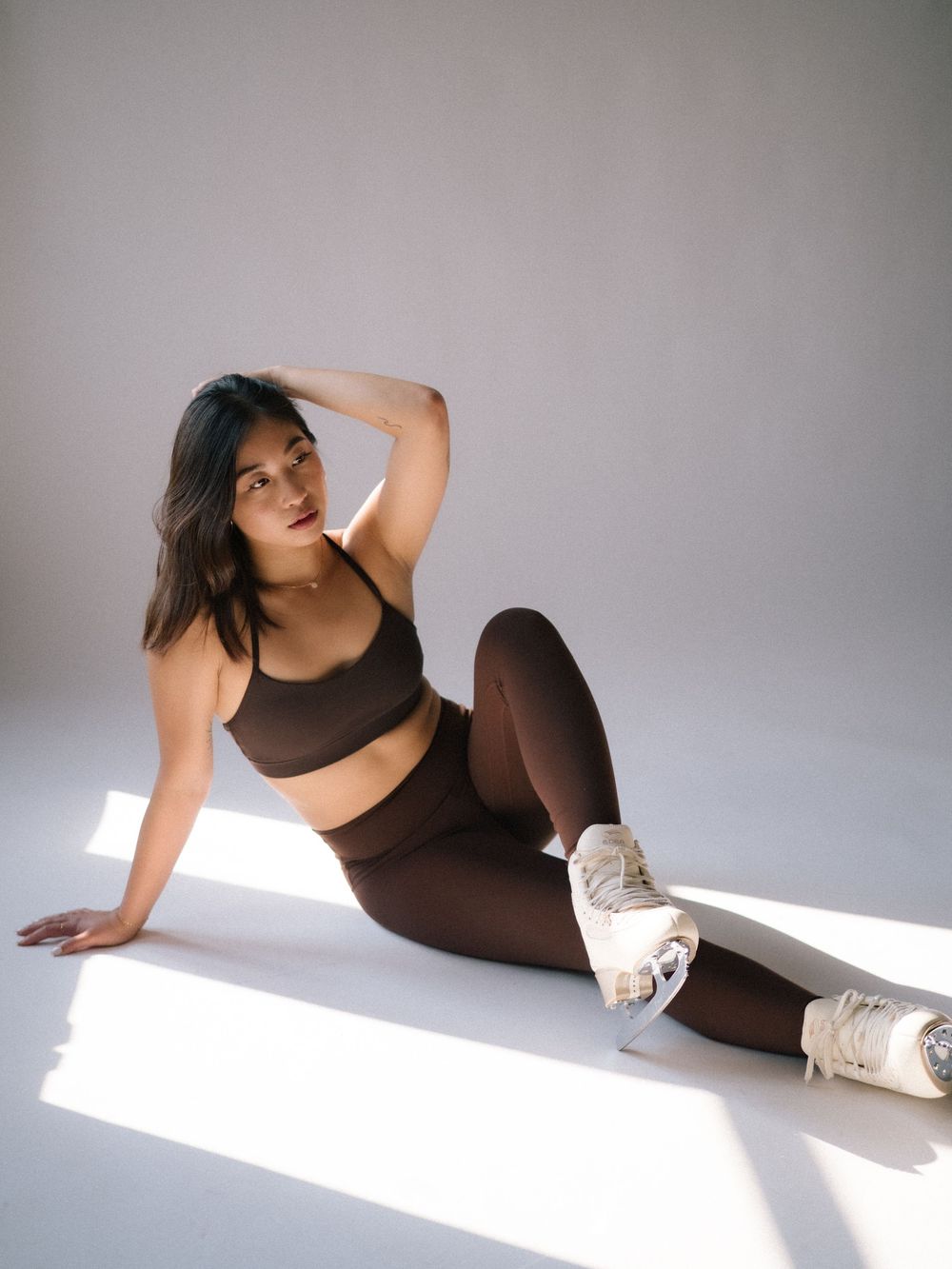 Athletic model poses in brown activewear set and white sneakers on minimalist white studio floor with dramatic lighting.