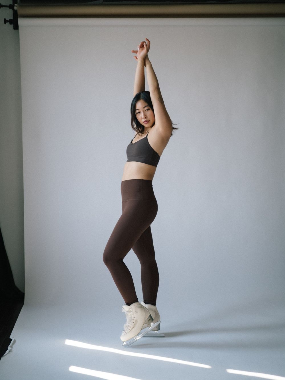 Athletic figure in brown leggings and black sports bra poses gracefully against white backdrop in dance-like stance.