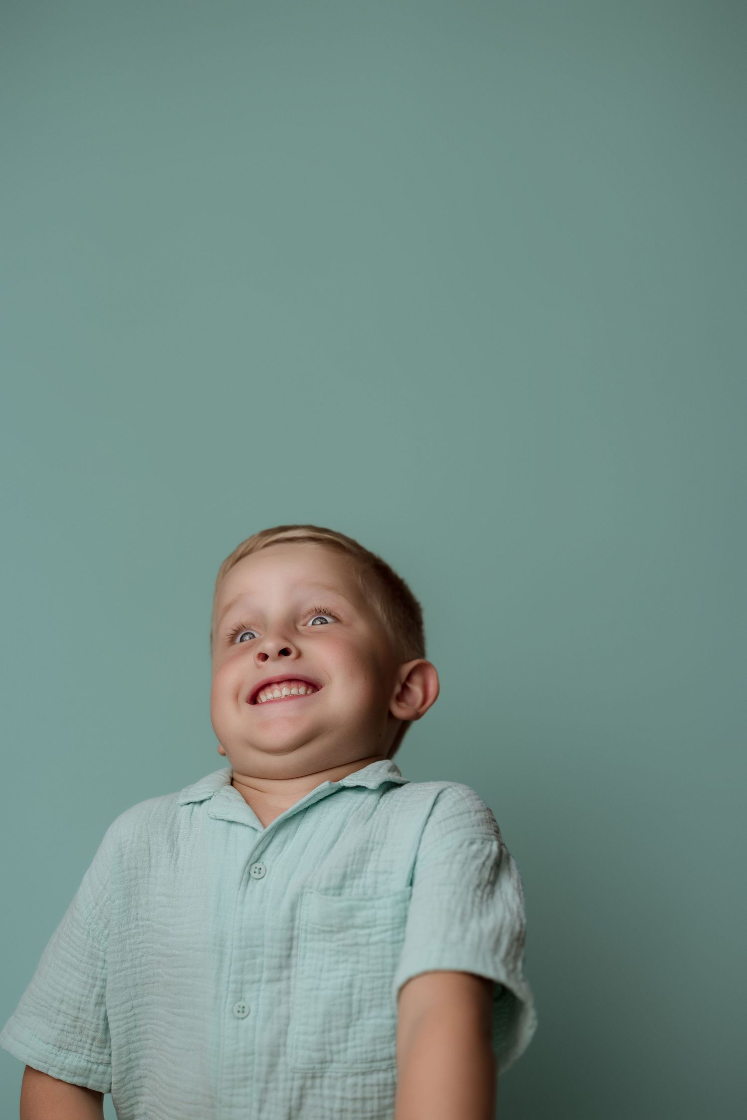 A child wearing a light mint green shirt looks upward with an excited expression against a teal background.
