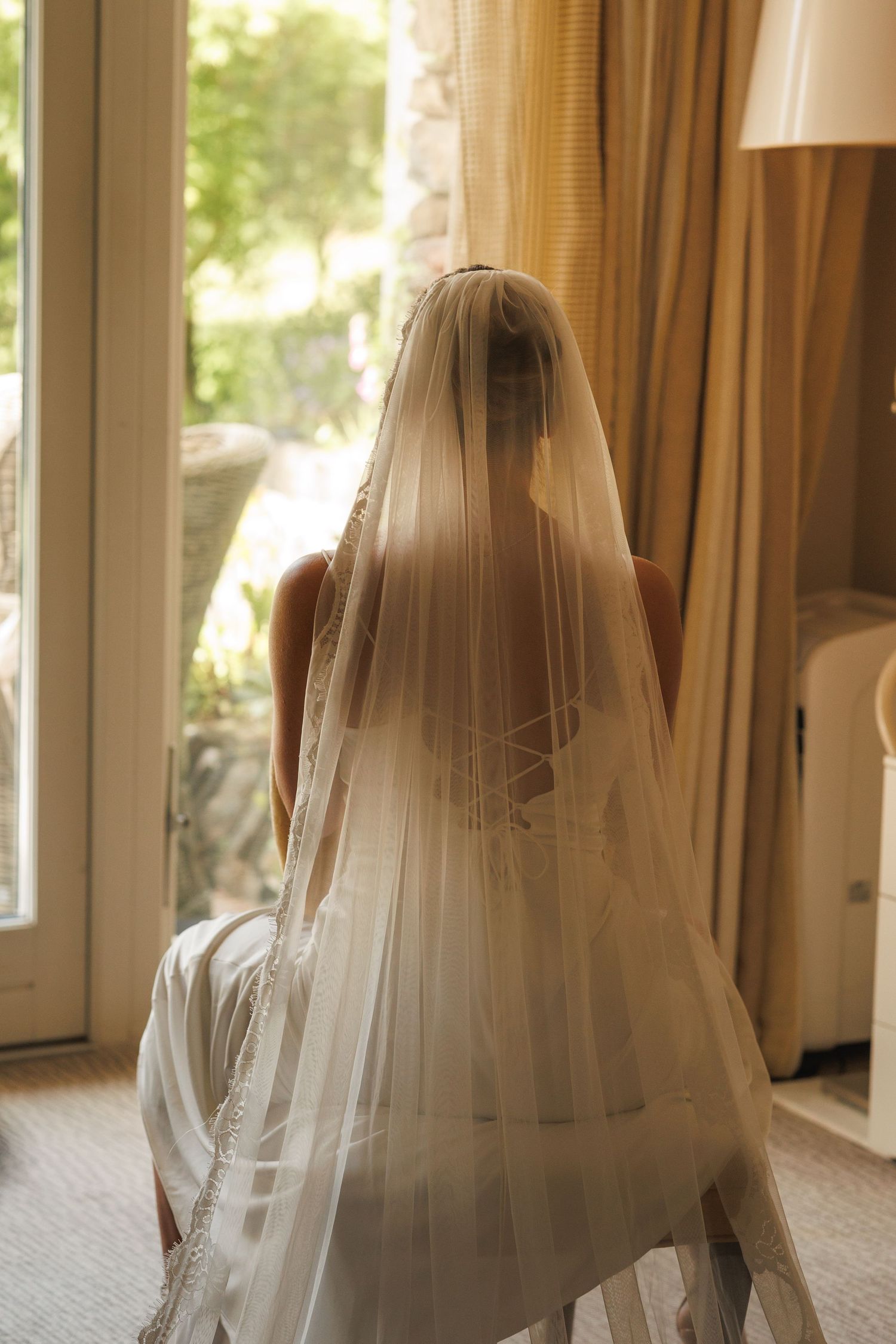 Ethereal backlit wedding veil silhouette captured through sunny window with sheer curtains.