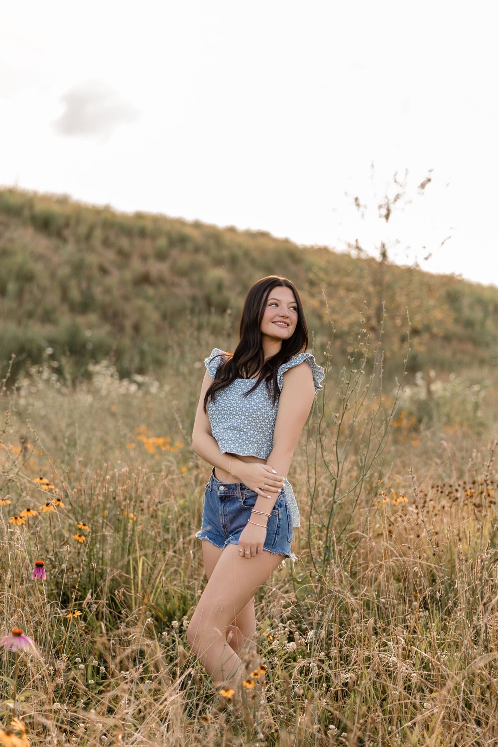 A young woman in denim shorts and polka dot top poses in a golden wildflower meadow during sunset.