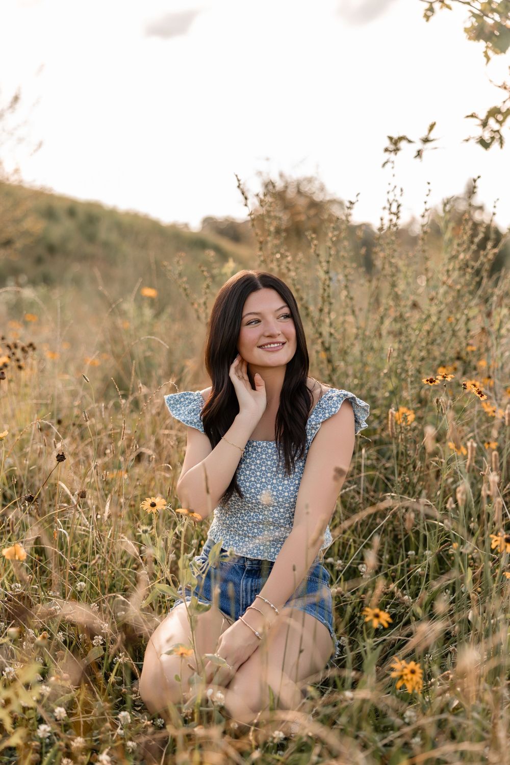 A person sits in a golden wildflower meadow at sunset wearing a blue floral top and denim.
