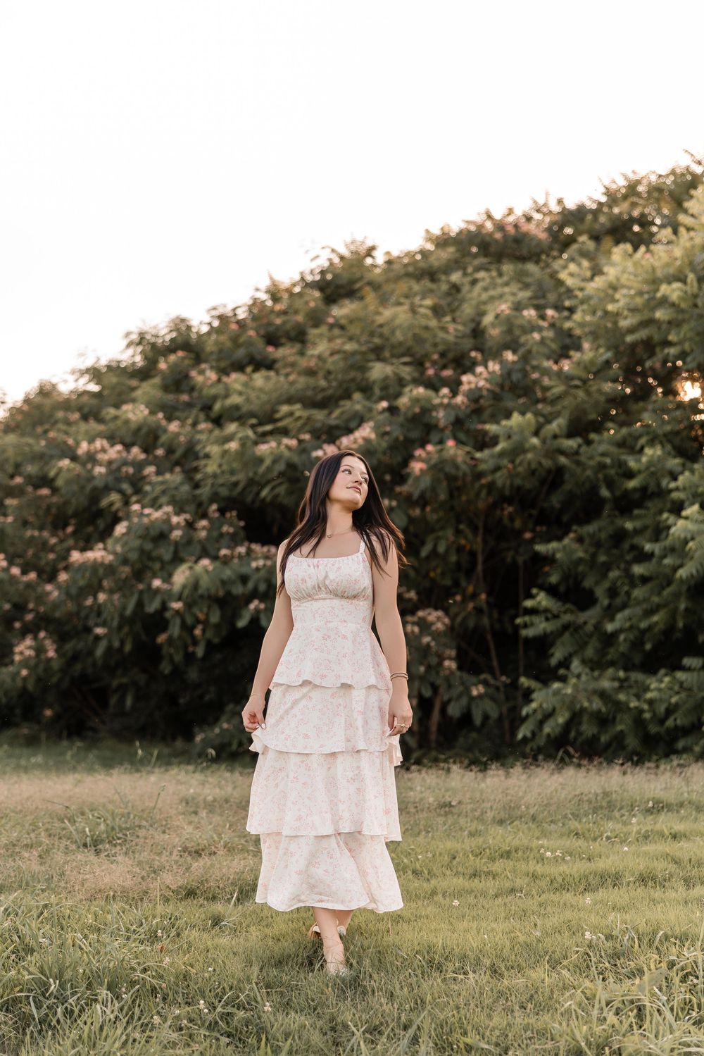 A person in a flowing white dress twirls gracefully in a grassy field with flowering bushes in the background.