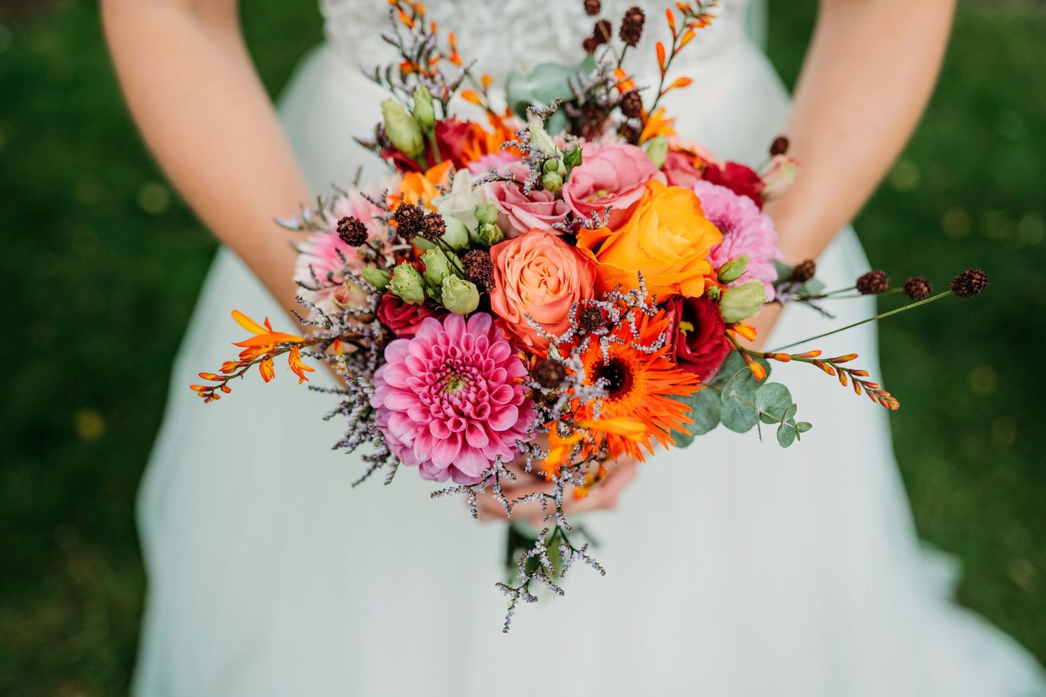 Vibrant autumn wedding bouquet with coral roses, orange ranunculus, and pink blooms held in front of sage green dress.