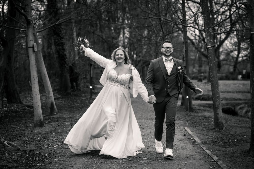 Ein glückliches Brautpaar spaziert Hand in Hand auf einem Waldweg – die Braut in einem fließenden Kleid, strahlend vor Freude,