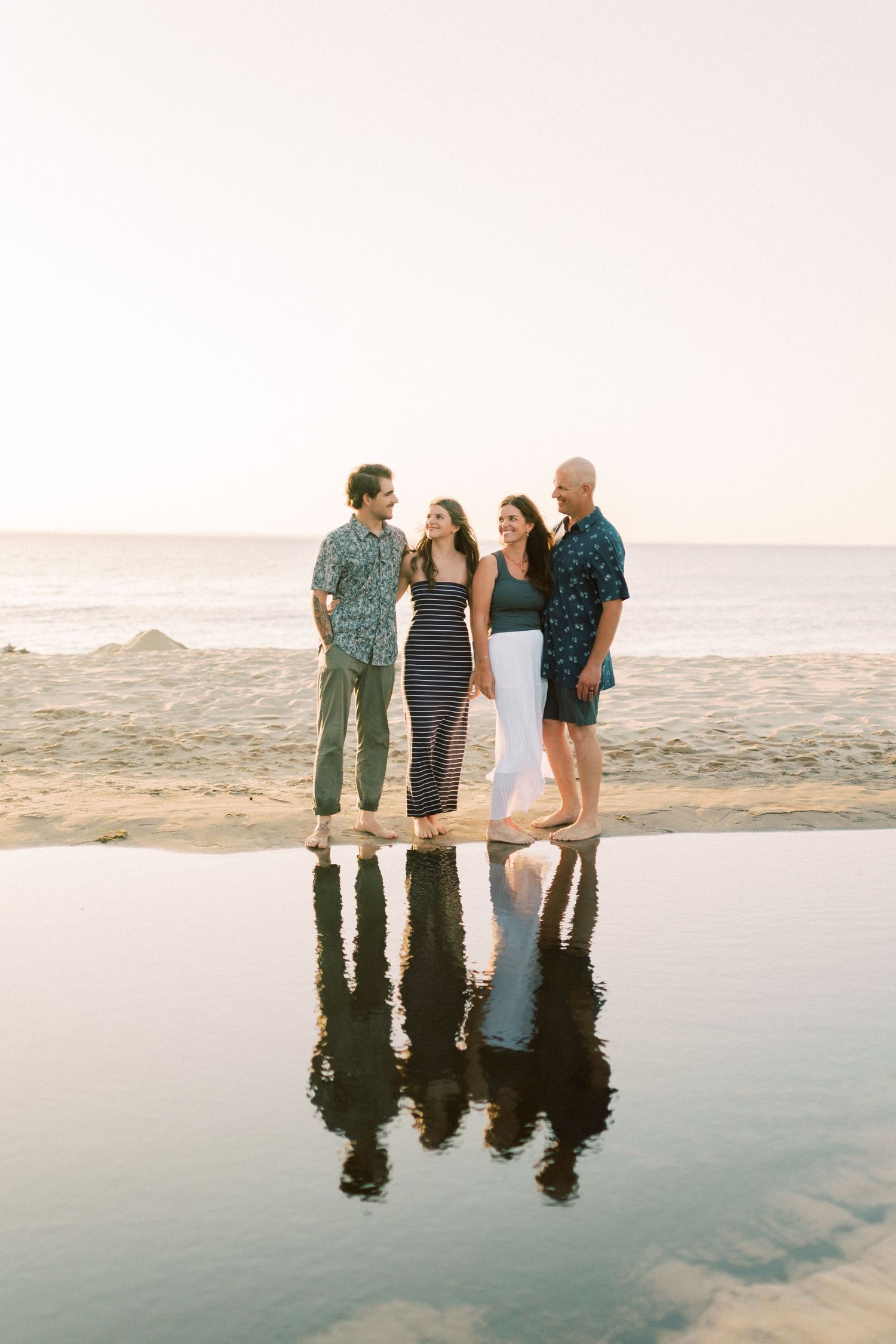 Two couples stand together smiling on a sandy beach at sunset with ocean waves and mountains in the background.