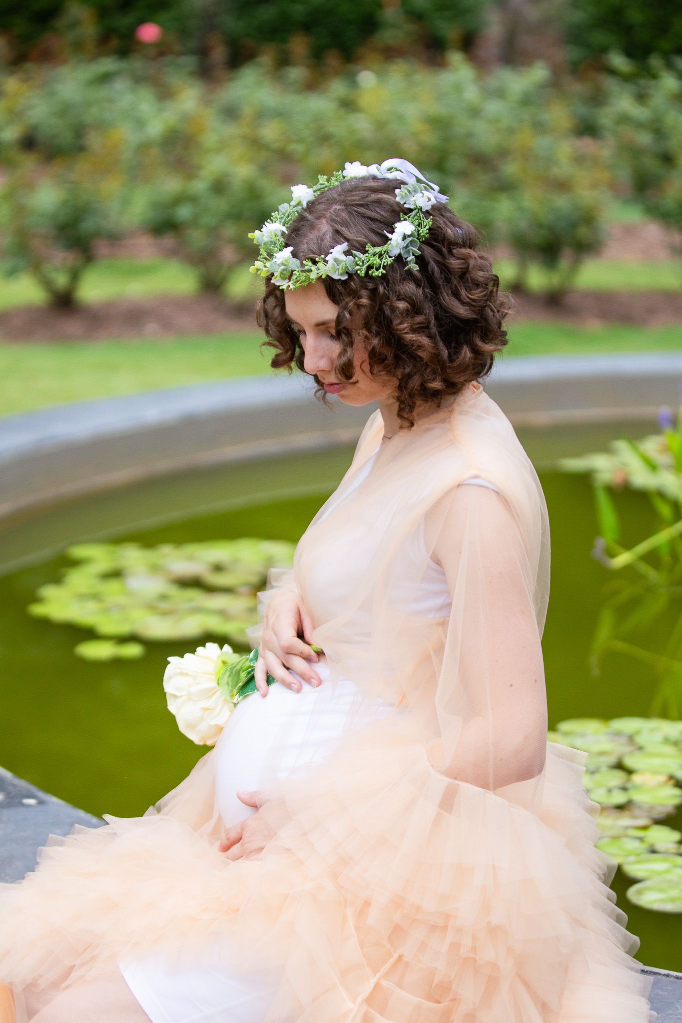 A woman in a blush-colored tulle gown poses near a lily pond during a romantic outdoor photo session.