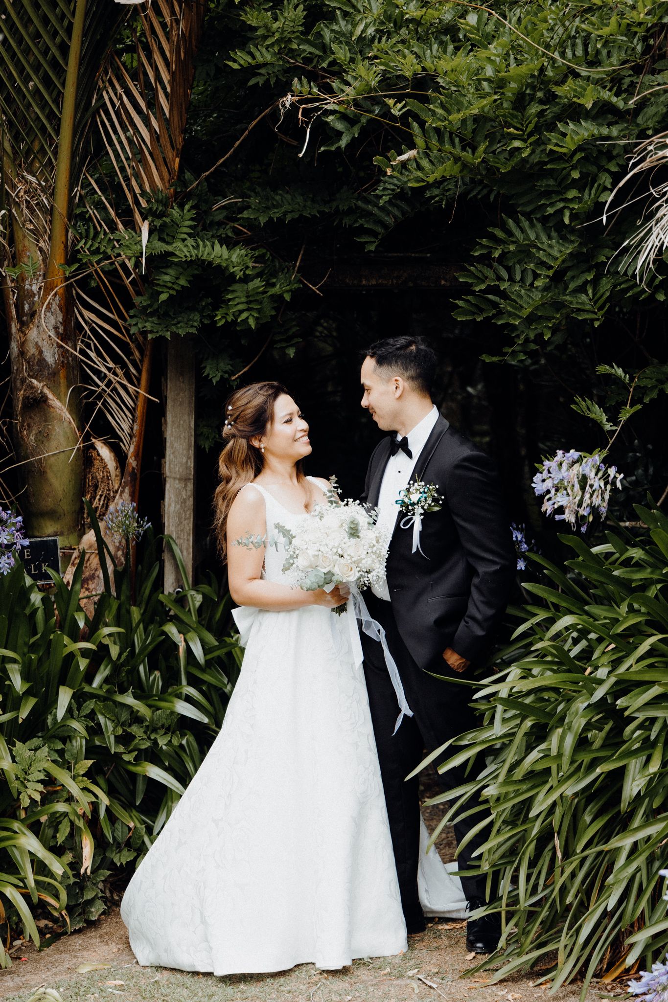 A wedding couple stands together among lush tropical foliage captured by Apertured Media Auckland Wedding Photographer.