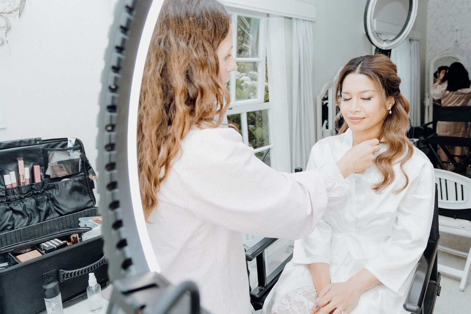 A makeup artist applies beauty products to a bride wearing a white robe captured by Apertured Media Auckland Wedding Photographer.