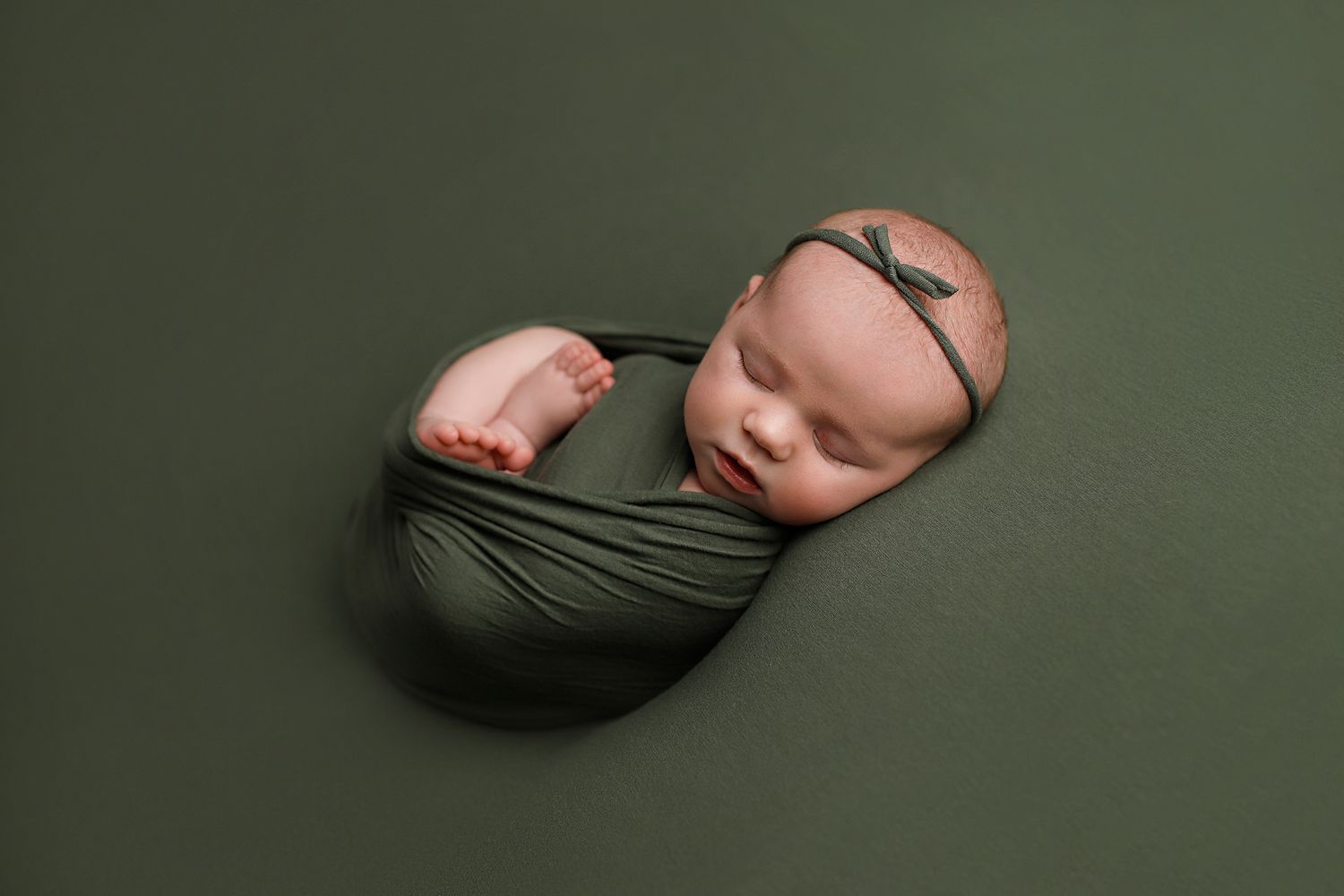 Newborn baby wrapped in olive green swaddle sleeping peacefully on matching green backdrop. Utah newborn photographer