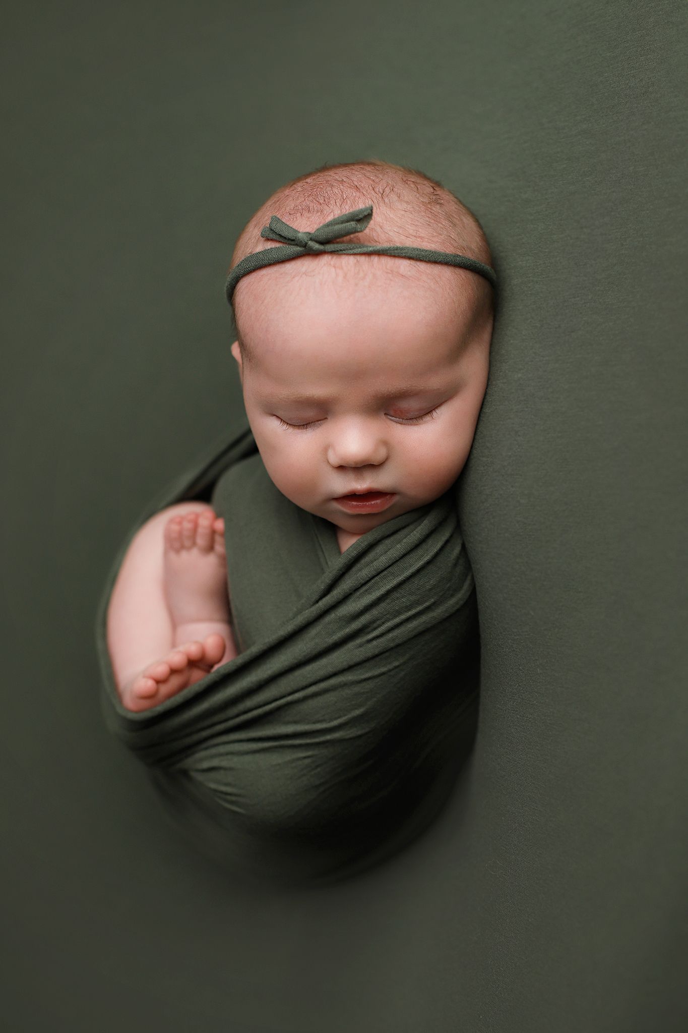 A sleeping newborn wrapped in olive green fabric against a dark green background wearing a matching bow headband. Orem newborn photography