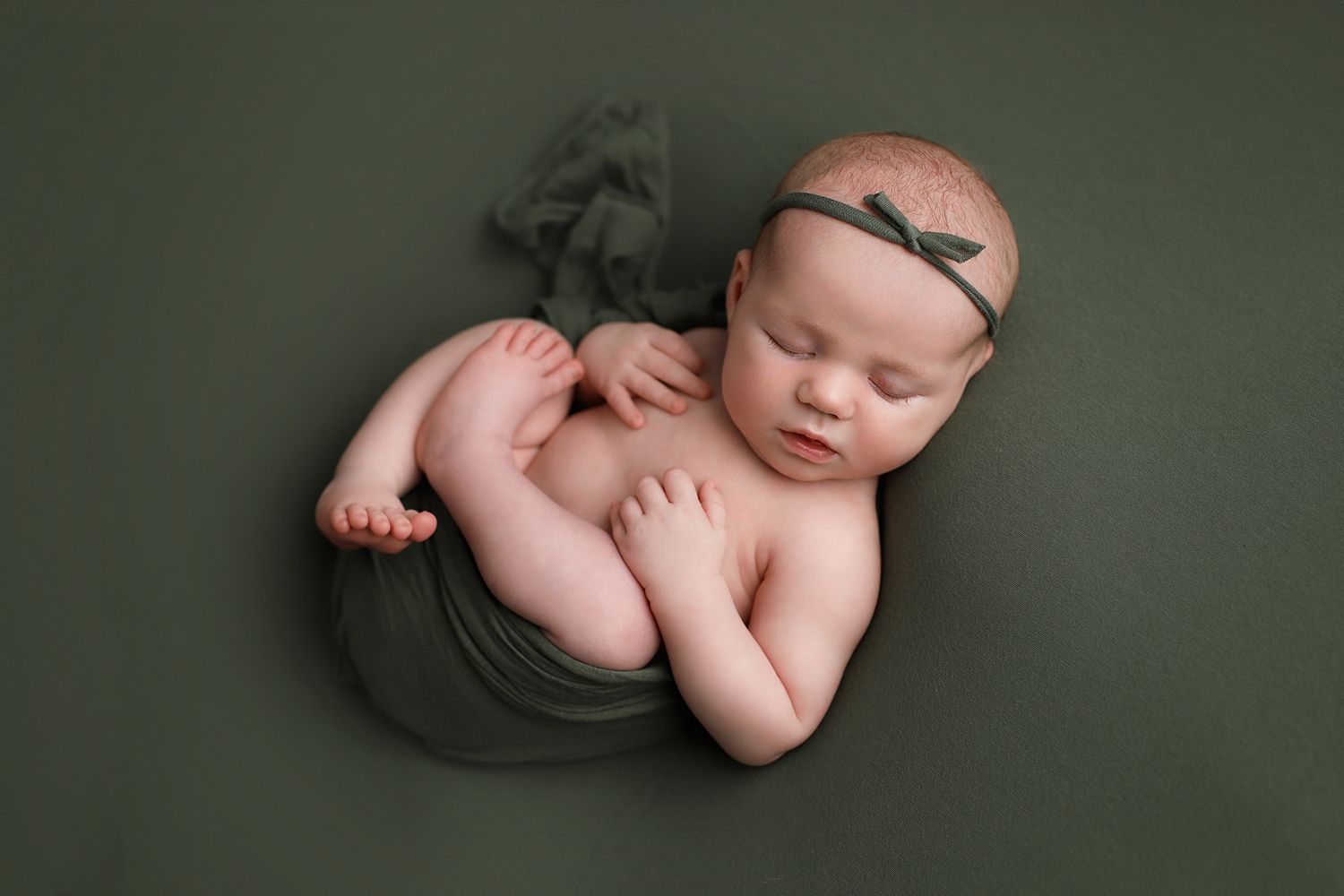 Peaceful sleeping newborn wrapped in olive green fabric on matching dark green background with delicate headband. Orem newborn photography
