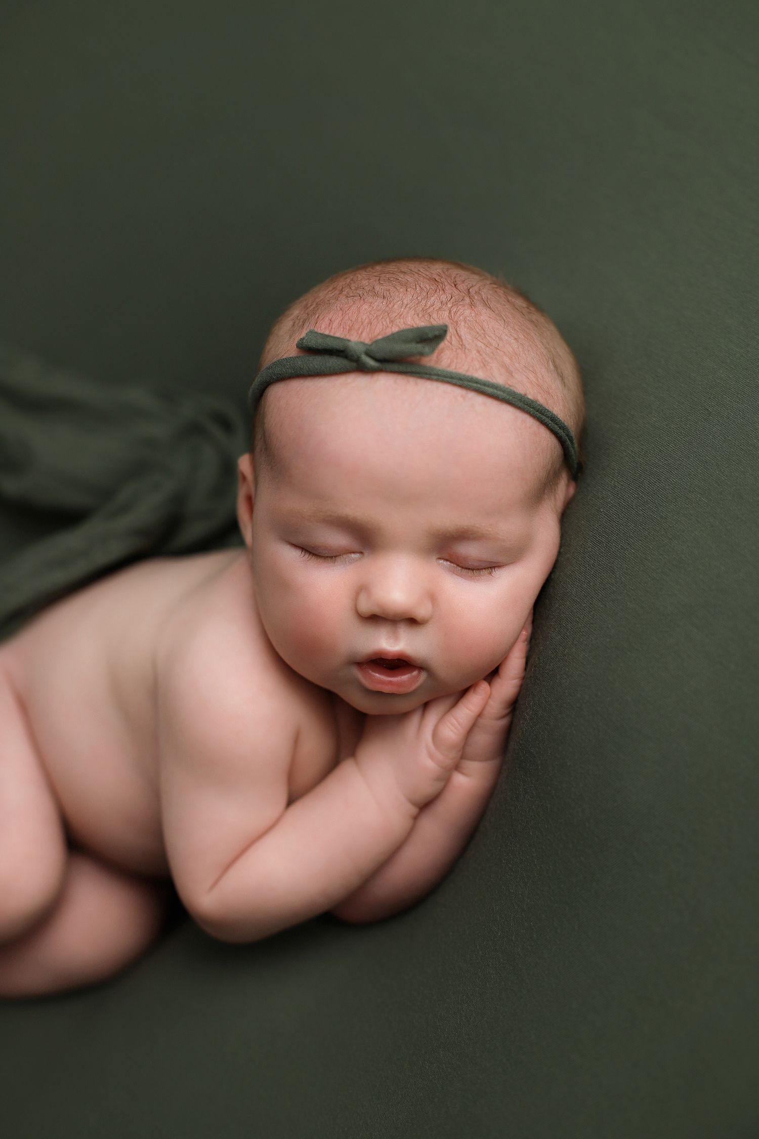Sleeping newborn baby wearing delicate headband rests peacefully on dark green backdrop during portrait session. Utah newborn photographer