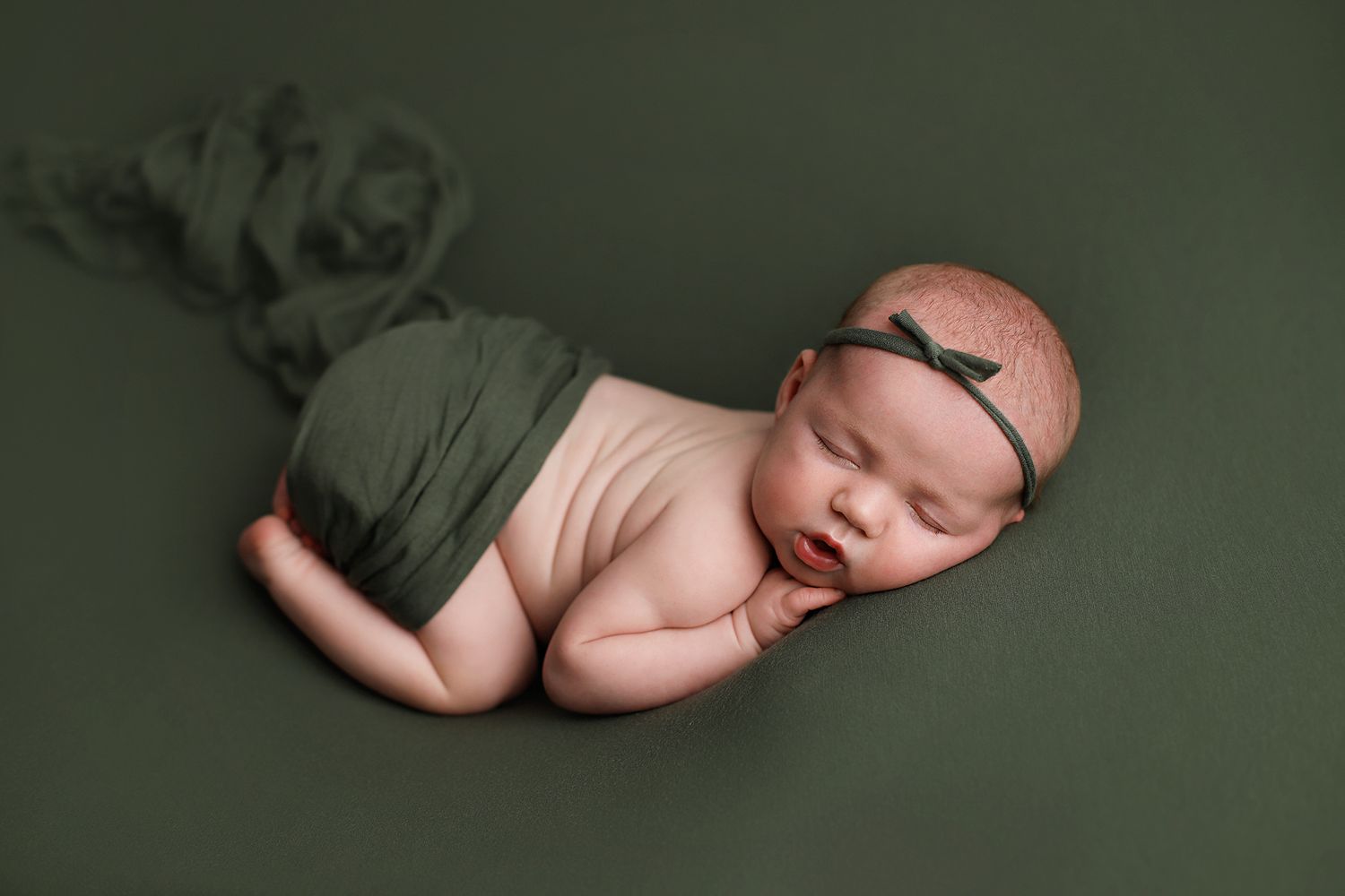 Newborn baby sleeps peacefully on dark green backdrop wearing matching headband during professional photo session. Orem newborn photography