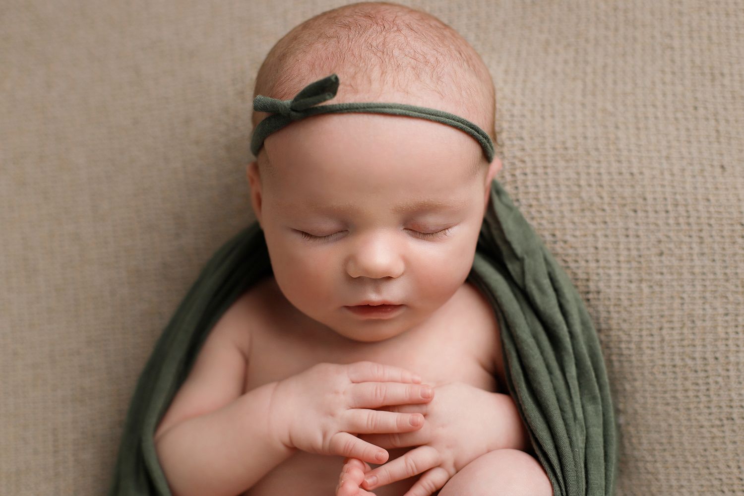 Peaceful sleeping newborn baby wrapped in olive green swaddle with matching headband resting on beige backdrop. Utah newborn photographer