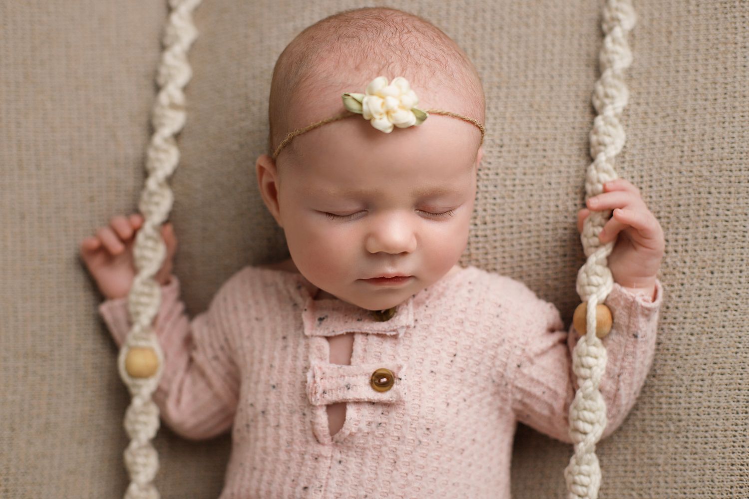 Baby in pink knitted sweater lying on beige fabric with decorative ropes and wearing a delicate floral headband. Utah newborn photographer