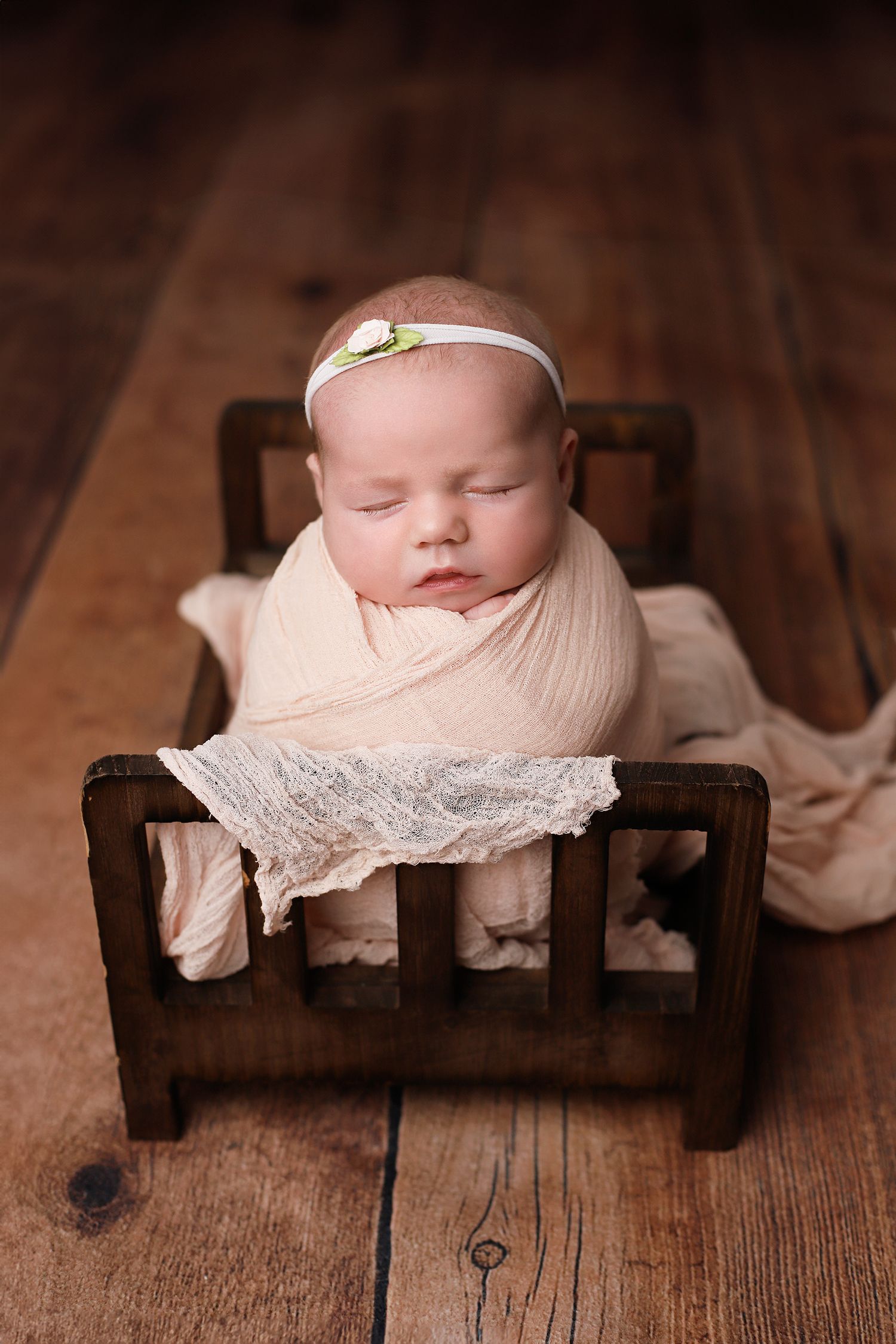 Sleeping newborn wrapped in pink swaddle lying in wooden prop bed with delicate headband on rustic wooden floor. Orem newborn photographer