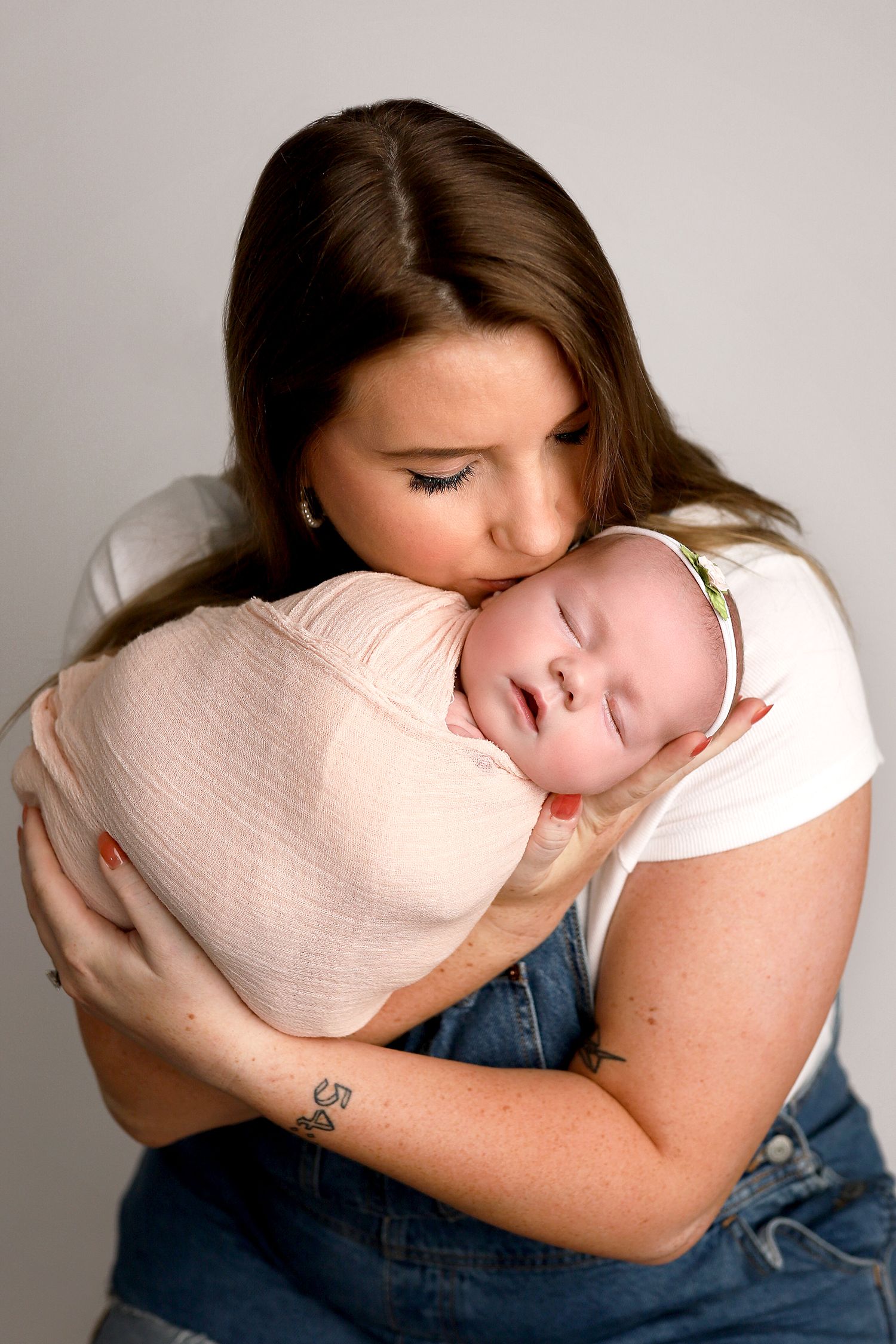 A woman in a white shirt and jeans tenderly embraces a sleeping newborn baby wrapped in soft pink swaddling. Orem newborn photography