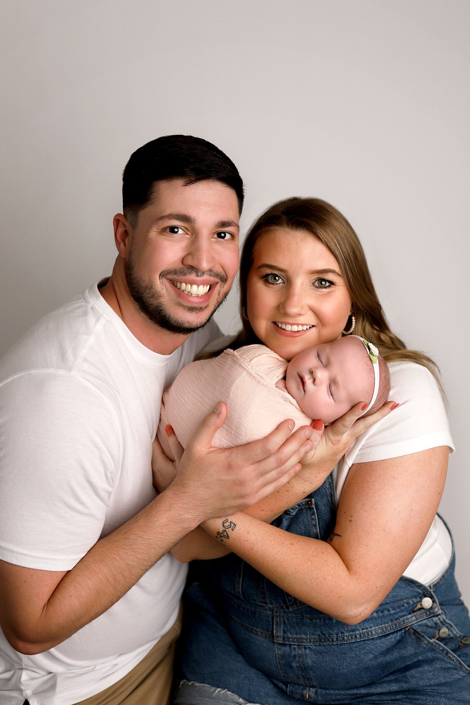 A couple in matching white shirts poses with their newborn baby while sitting against a neutral gray backdrop. Orem studio photography