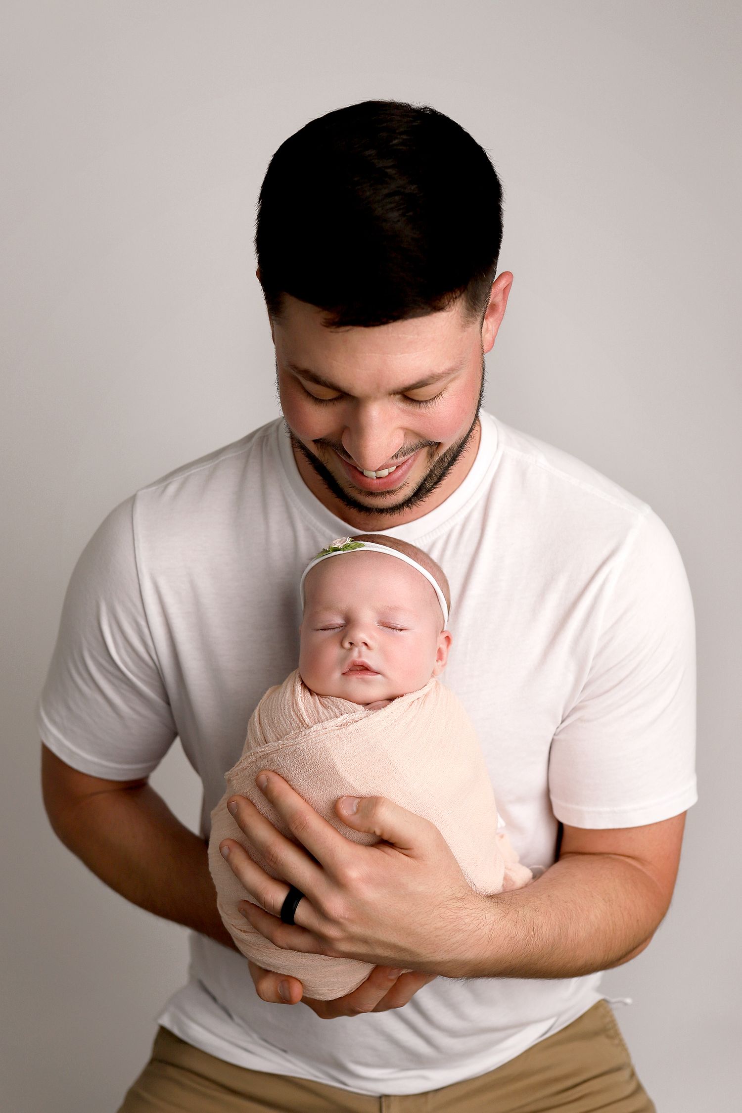 A loving parent wearing a white t-shirt holds and smiles down at a sleeping newborn baby wrapped in light beige fabric.