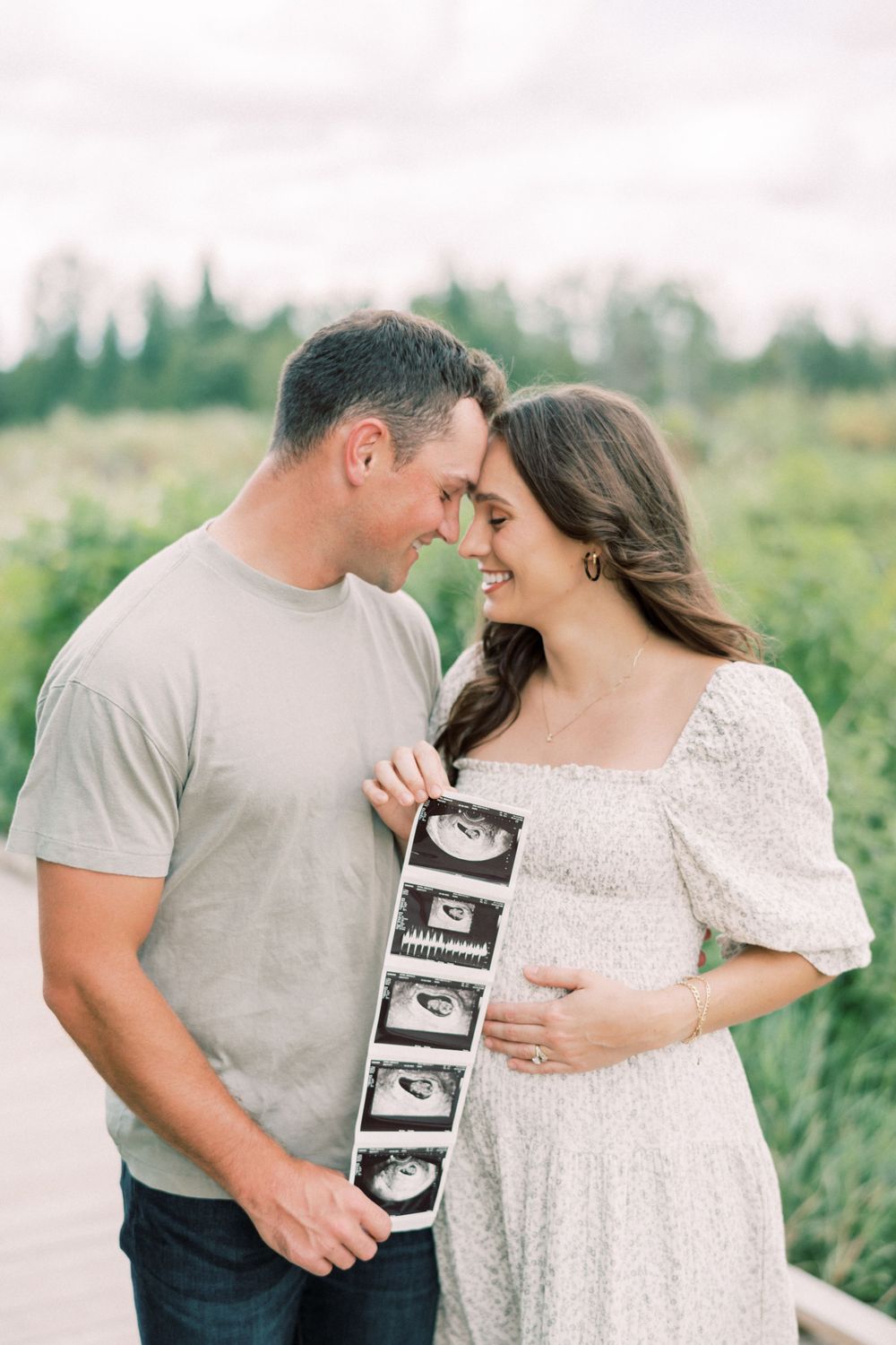 Expecting couple sharing intimate moment while holding ultrasound photos in nature setting.