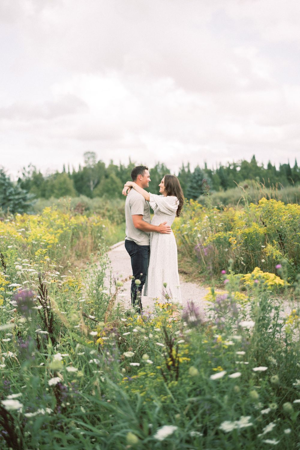 A romantic couple embraces on a nature trail surrounded by yellow wildflowers and greenery.