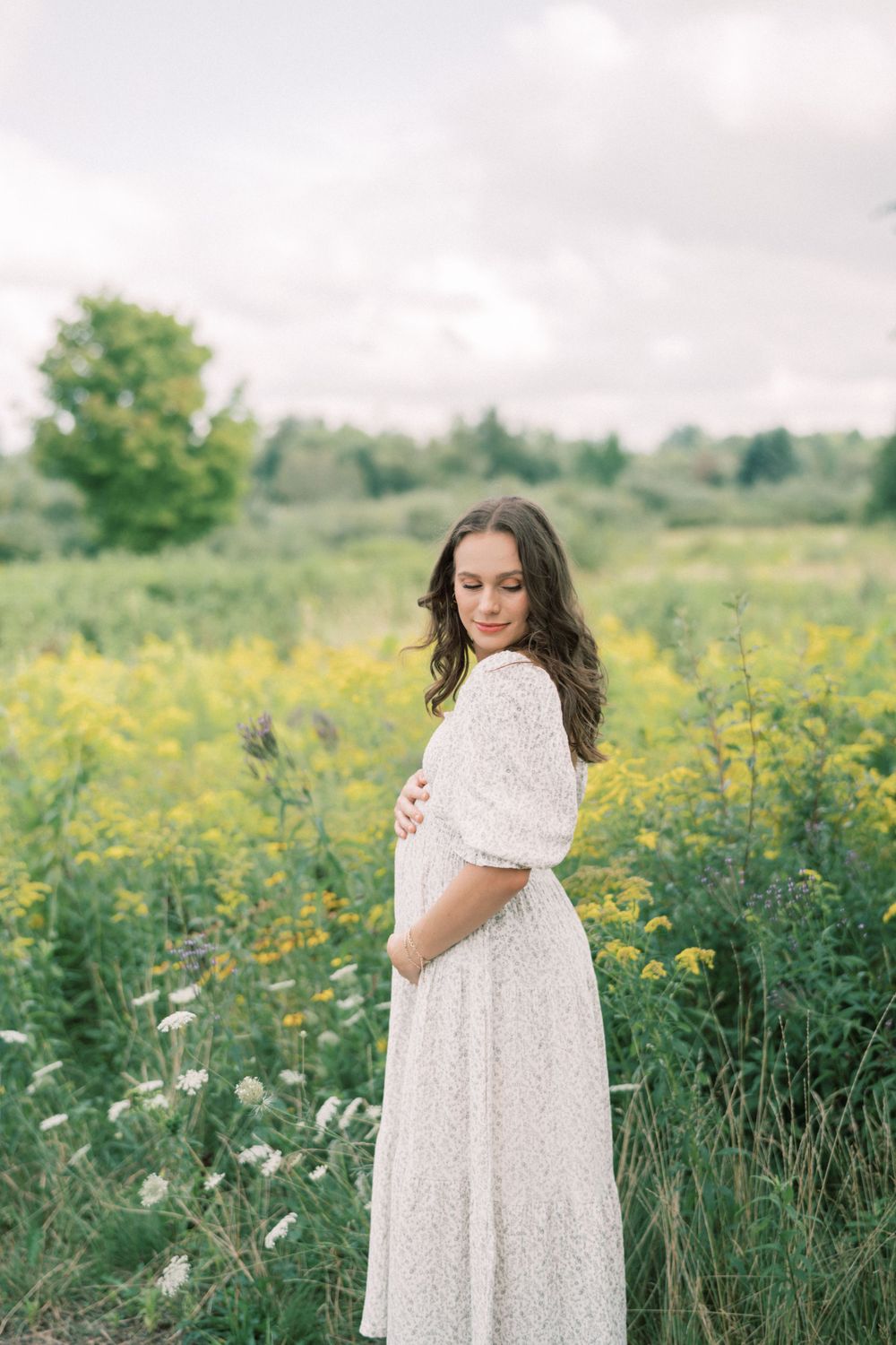 Woman in white lace dress poses in a meadow of yellow wildflowers under cloudy skies.