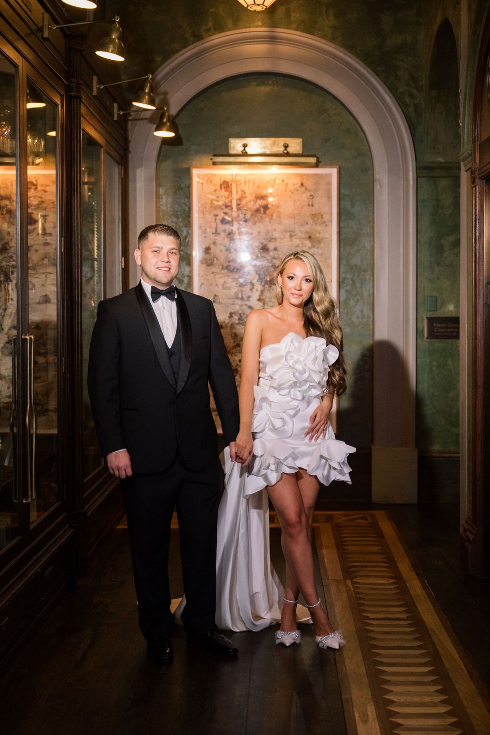Couple in formal attire standing in luxurious hallway with ornate arched architecture.