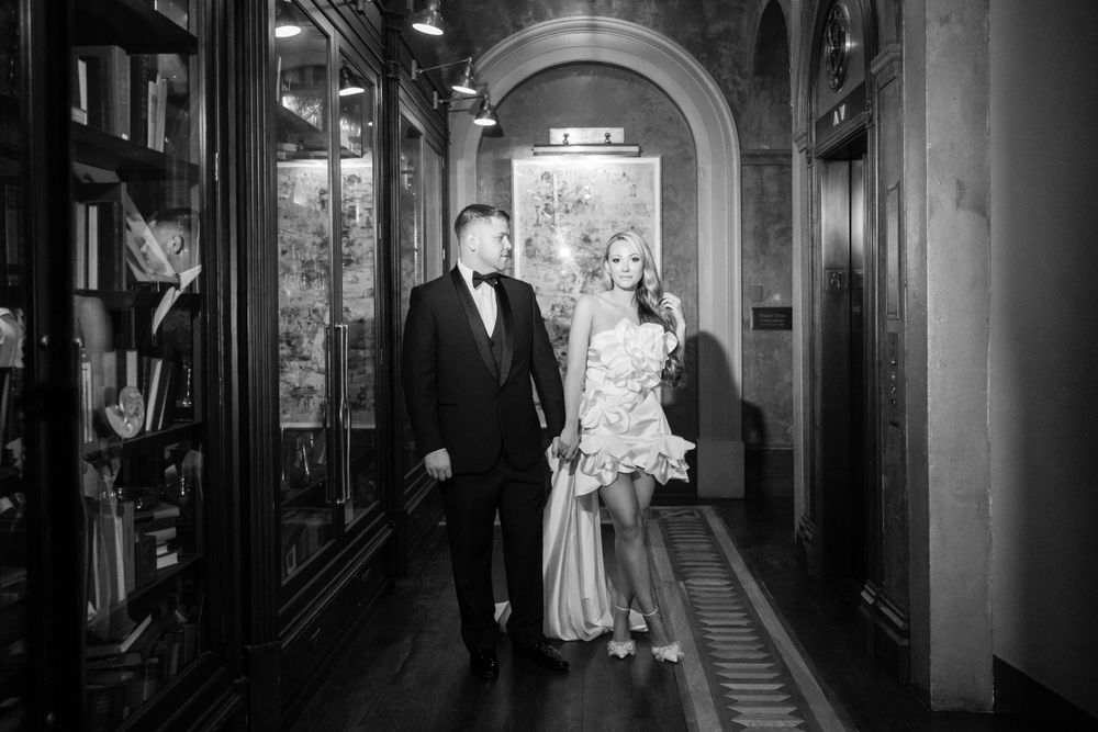 Black and white photo of couple in formal wear standing in historic hallway with arched architecture.