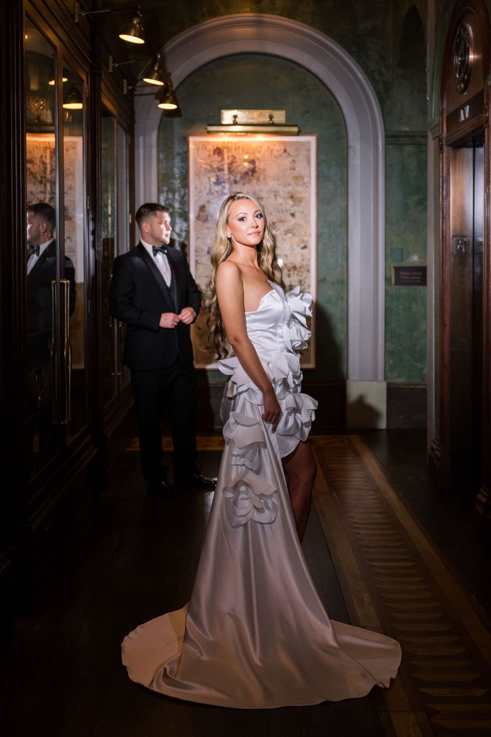 Glamorous formal portrait in an arched hallway showing a one-shoulder white gown with ruffled details.