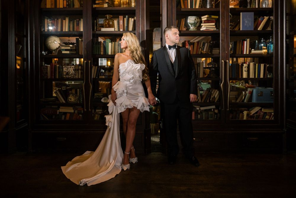 Elegant couple posed in formal attire against classic library backdrop with wooden bookshelves.