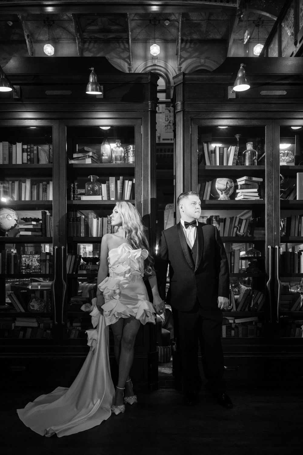 Formal couple posing in front of grand library bookshelves in black and white photography.