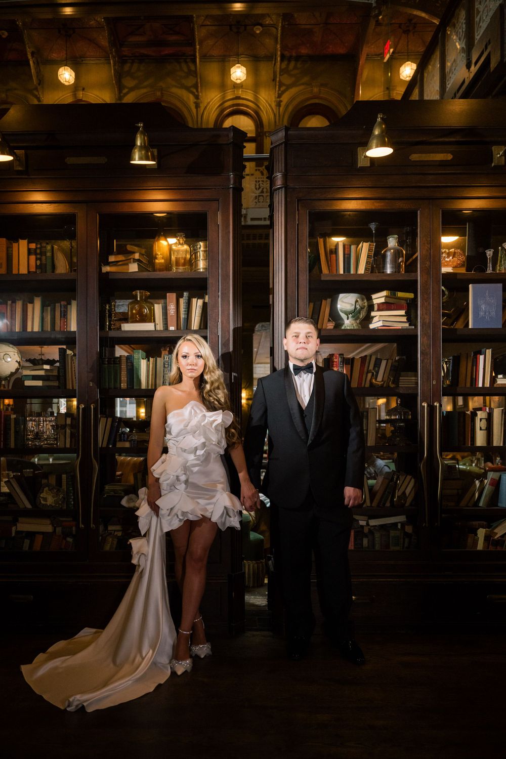 Stylish wedding portrait in library featuring contrast of white ruffled dress and classic black tuxedo.