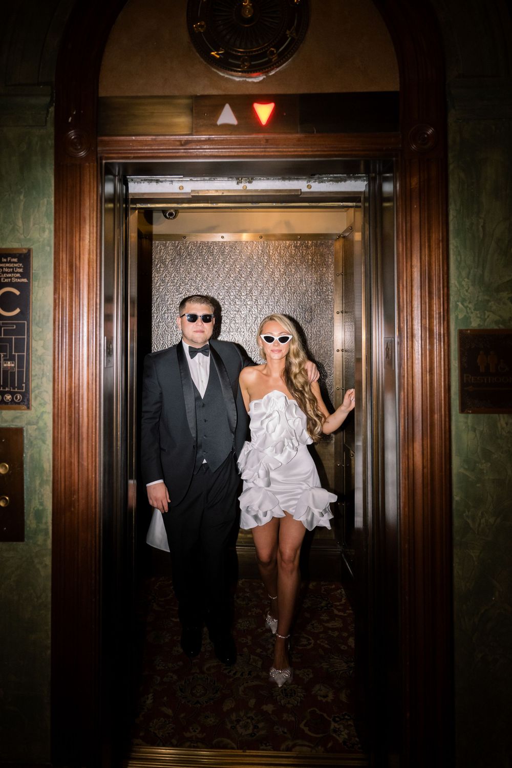 Stylish couple in formal wear posing in vintage elevator with patterned wallpaper.
