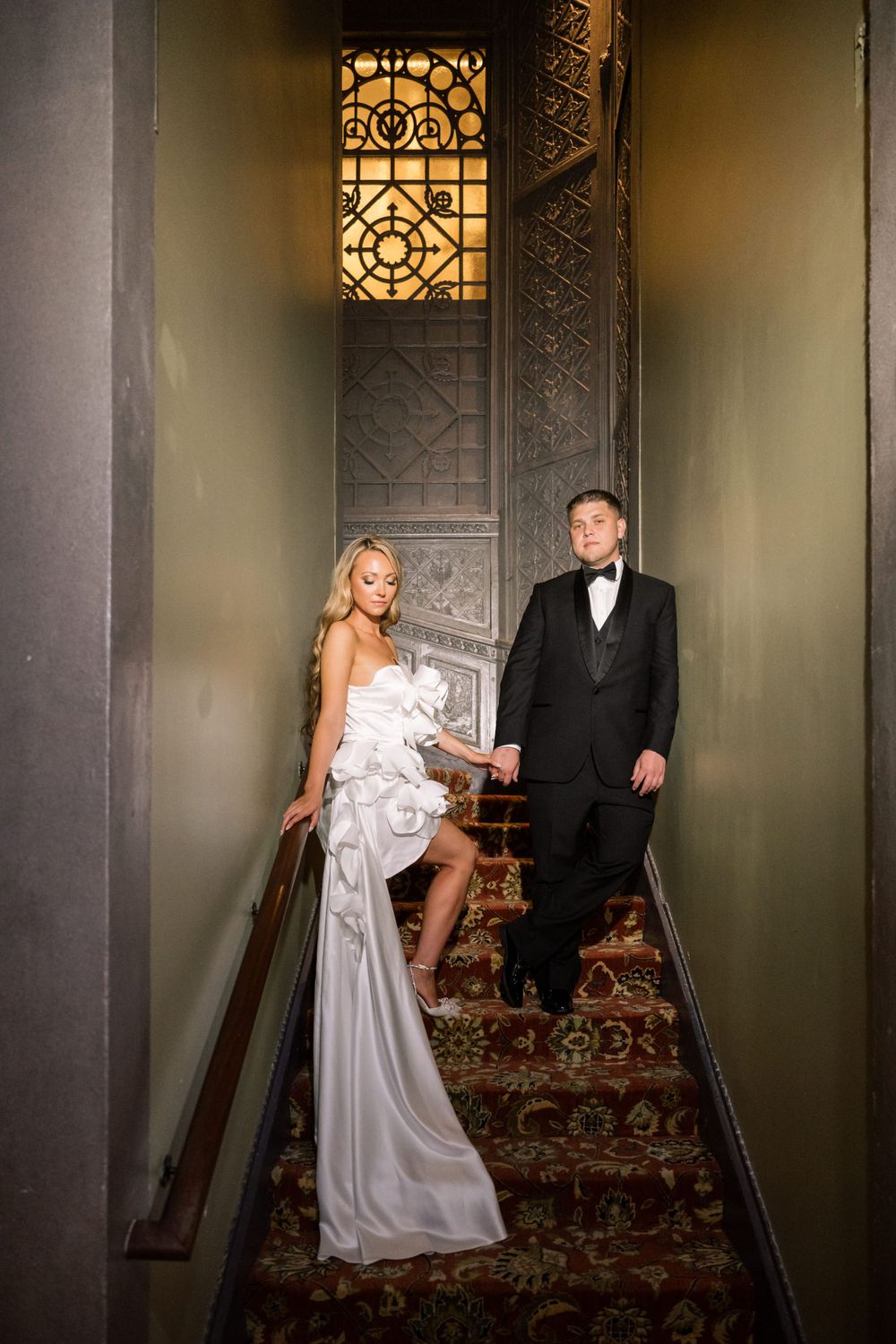 Wedding couple poses on ornate staircase with vintage stained glass window in historic building interior.