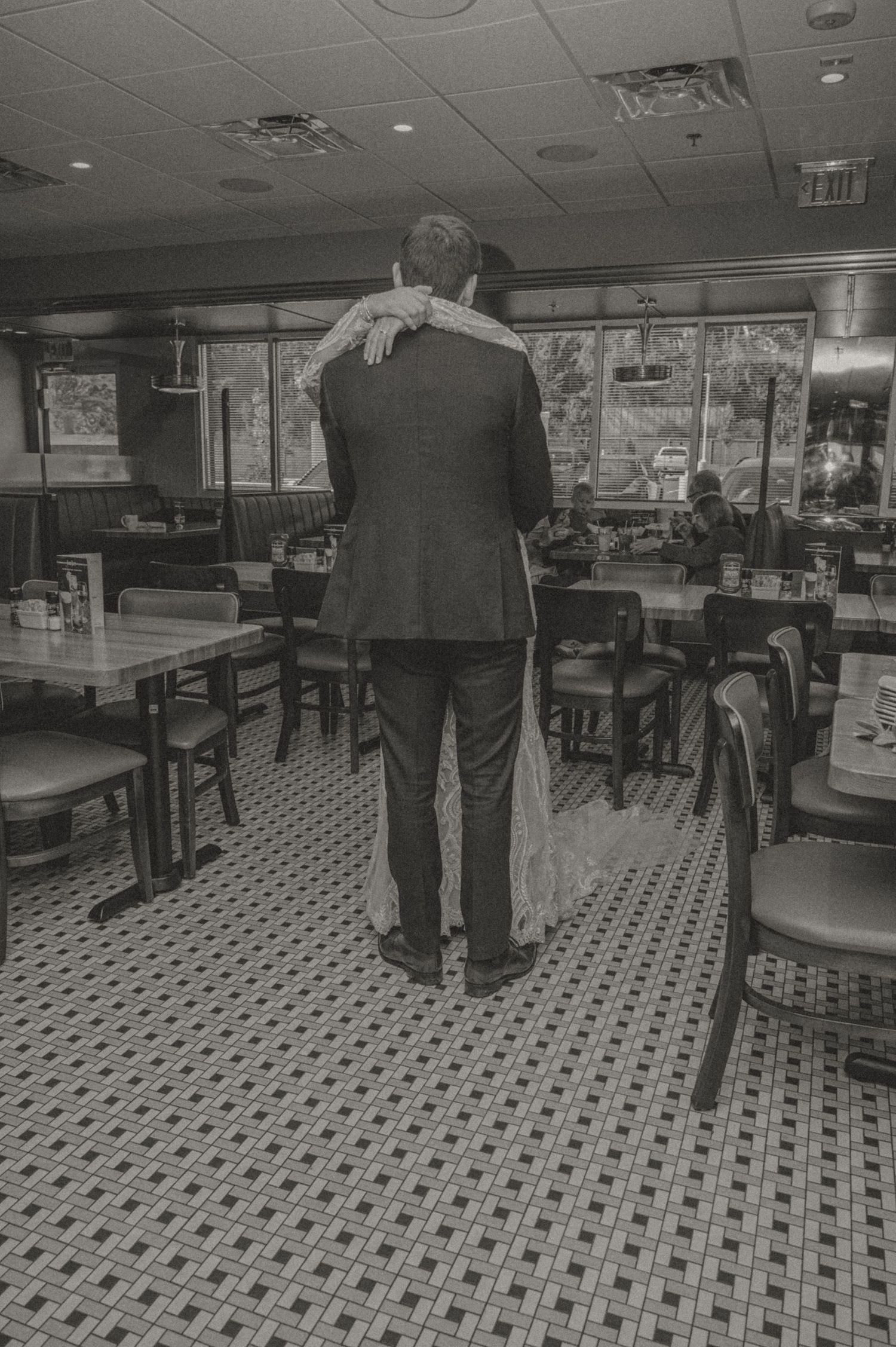 A black and white photo by Samantha Campbell Imagery of couple dancing in a diner with classic checkered floor tiles and booth seating.