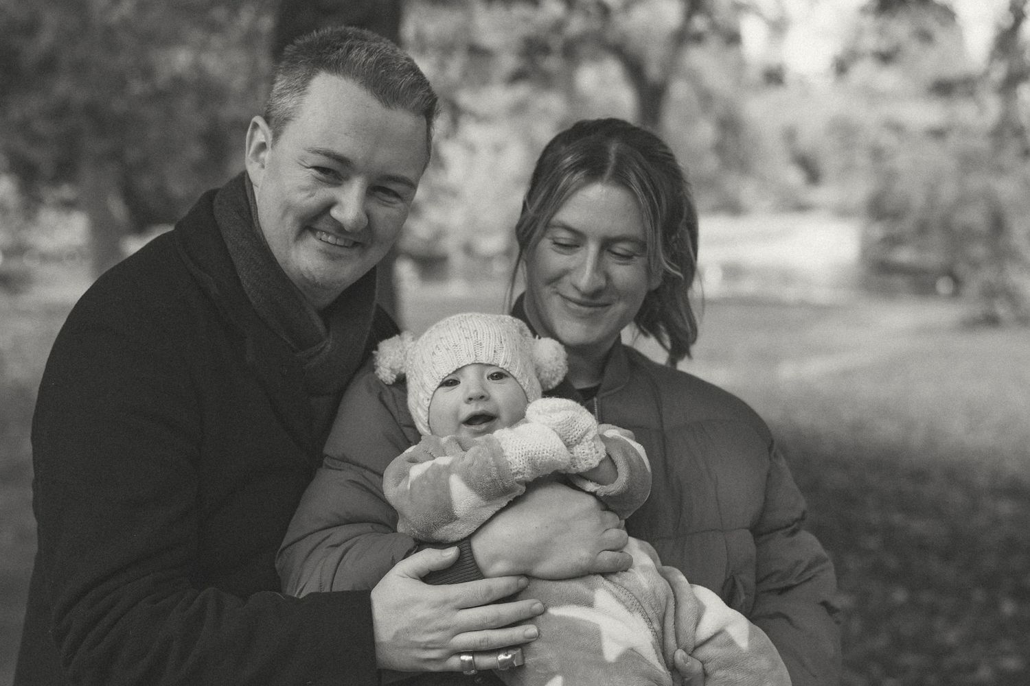 A black & white outdoor portrait of a couple holding their baby and smiling during their family photo session.