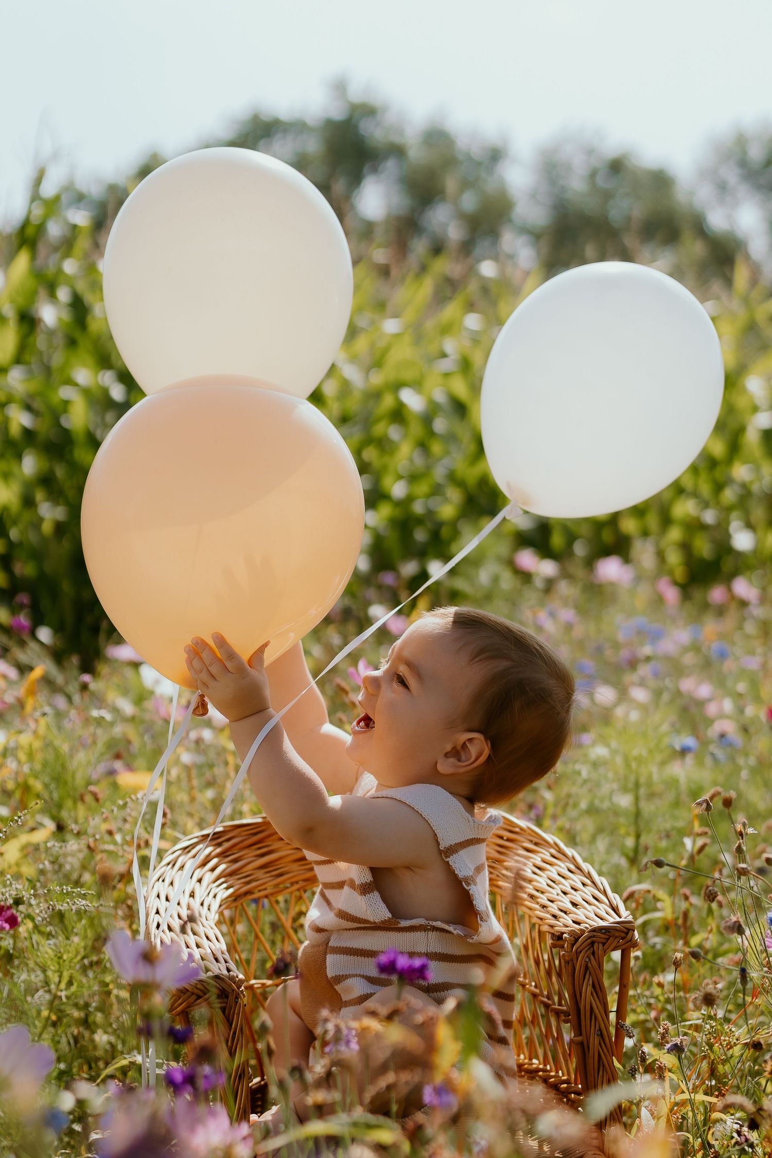 Happy toddler reaching for white and peach balloons while sitting in wildflower field.