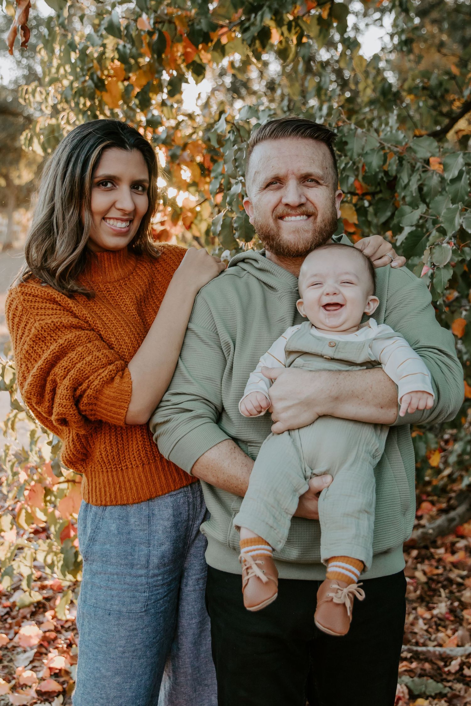 Happy family with baby poses together among autumn foliage at sunset.