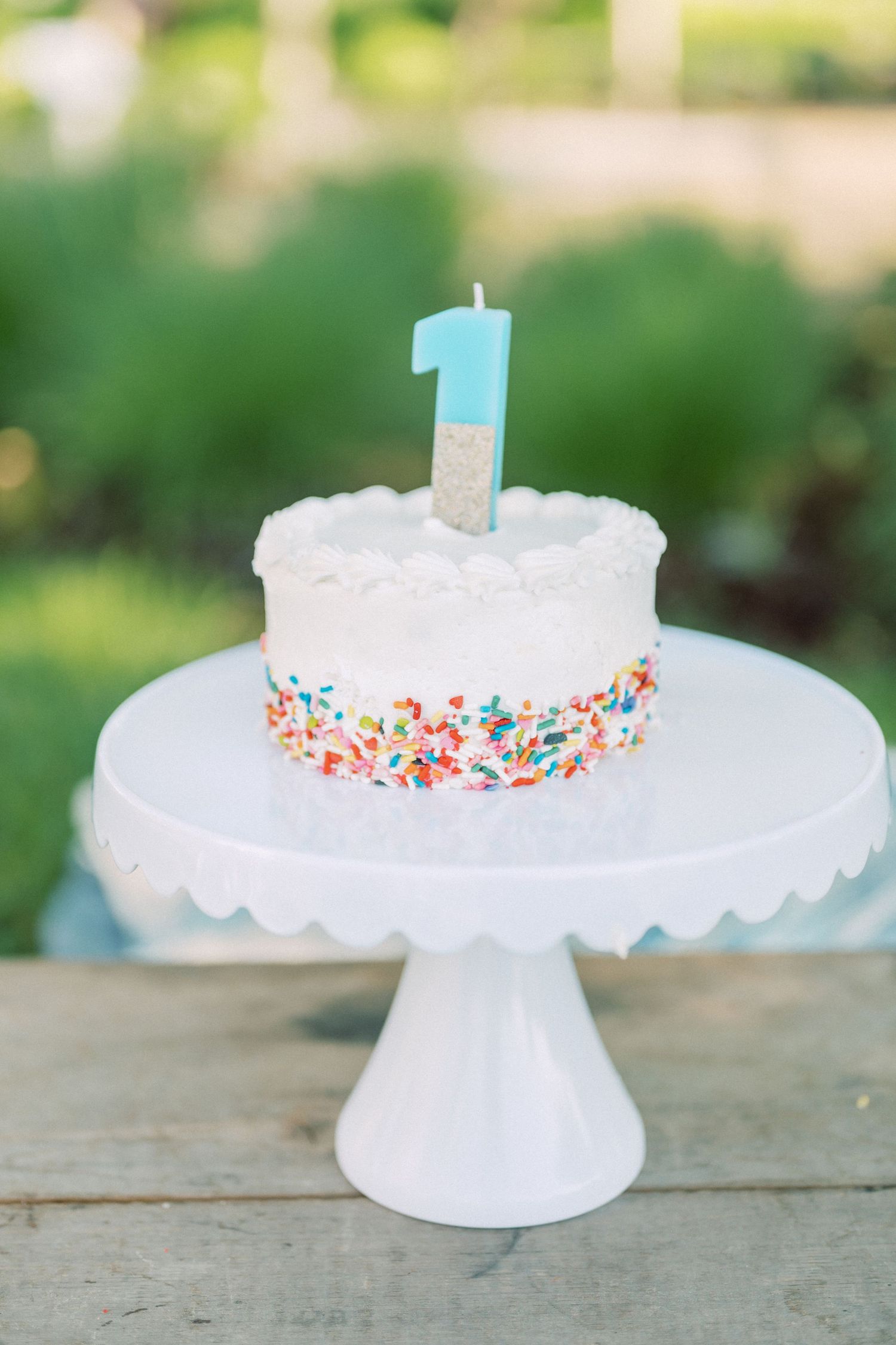 White birthday cake with blue number 1 candle and colorful sprinkles displayed on pedestal cake stand.