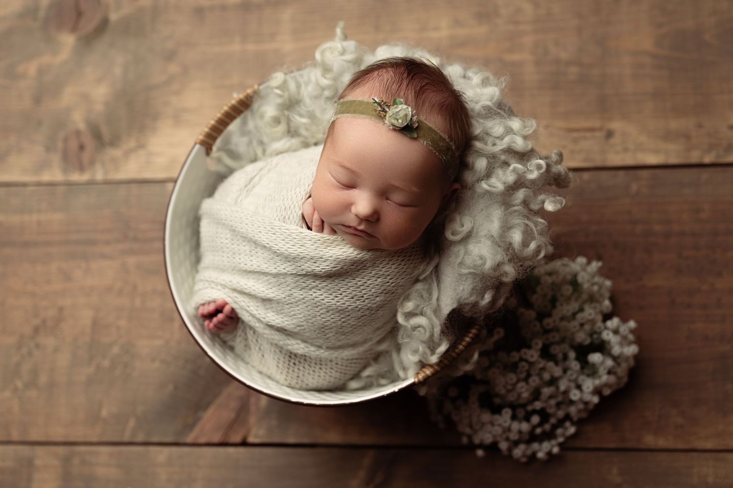 A peaceful sleeping newborn swaddled in cream fabric lies in a round bowl on rustic wooden planks with white flowers nearby.