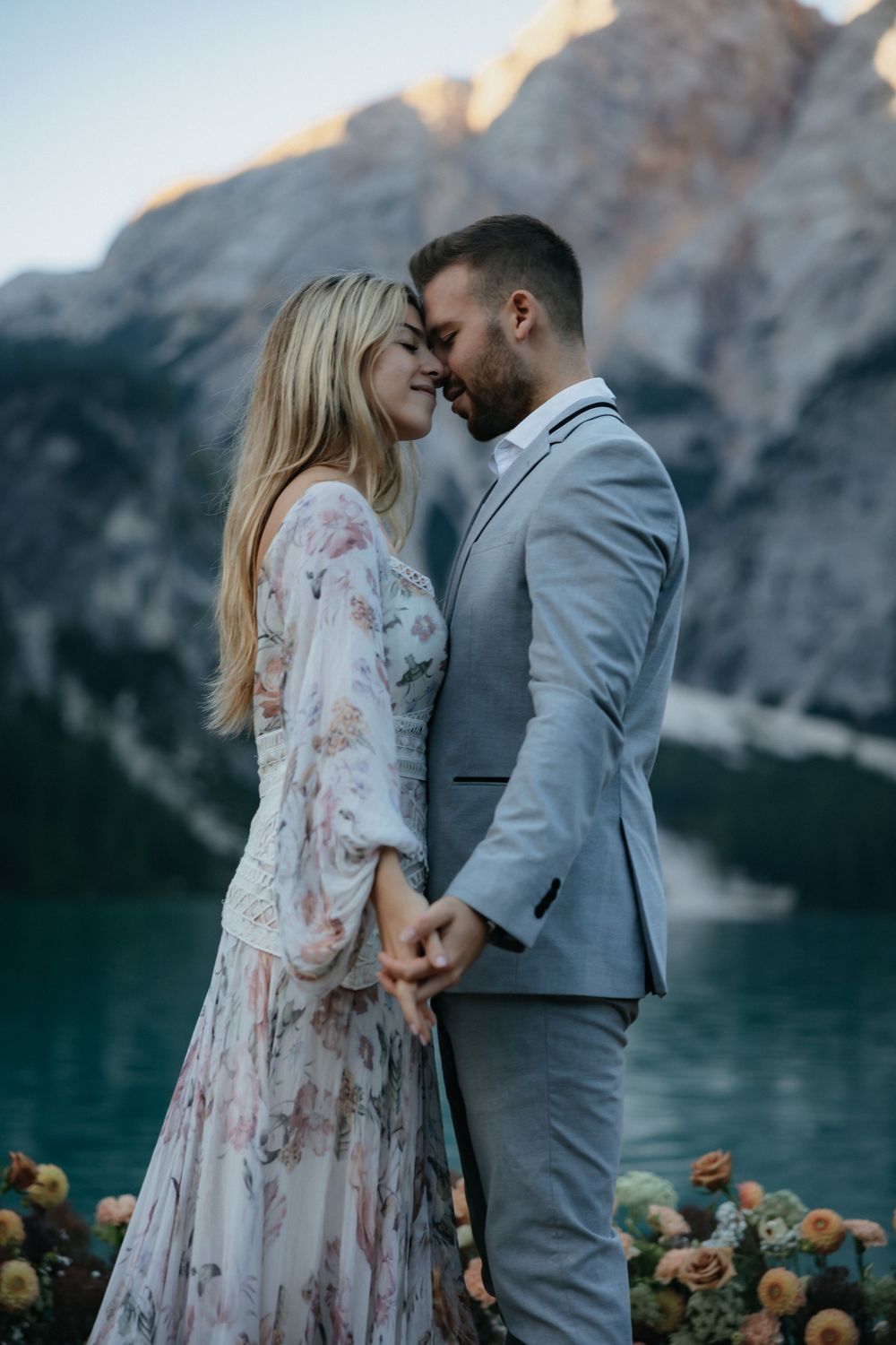 Romantic couple shares an intimate moment by a mountain lake at dusk in floral dress and gray suit.
