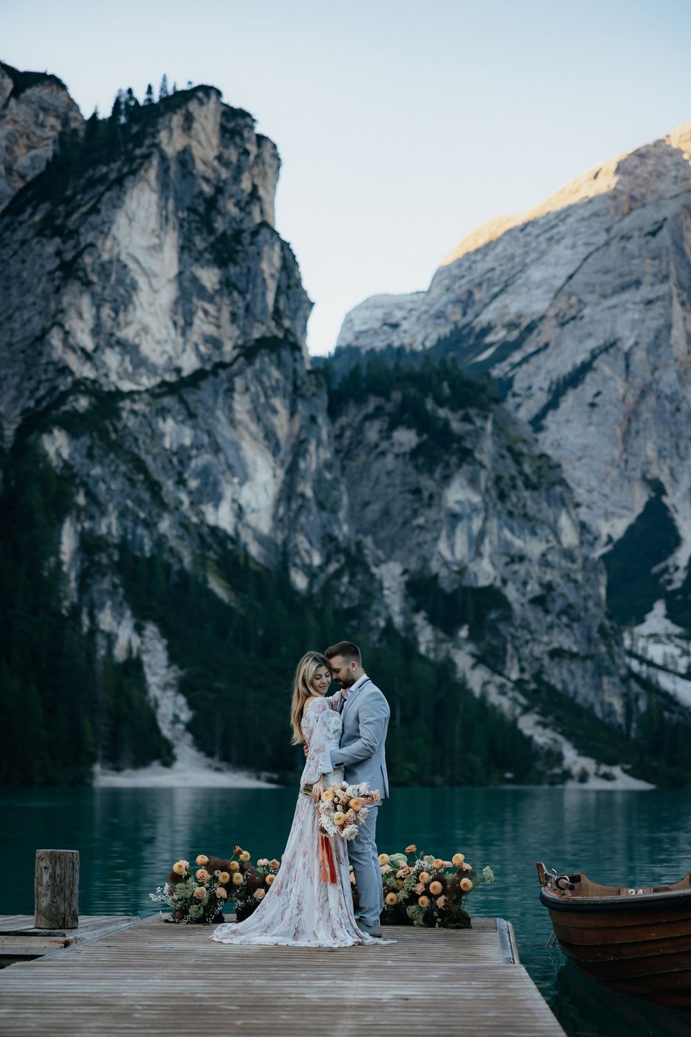Wedding ceremony setup on wooden dock overlooking dramatic mountain lake with wooden boats in background.