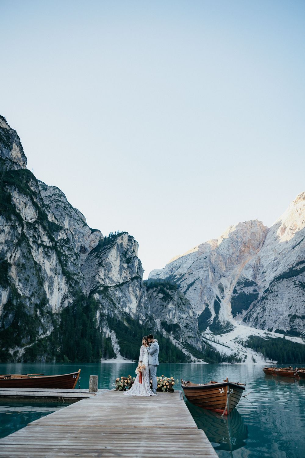 Wedding couple stands on wooden pier overlooking majestic Dolomites mountains and lake with wooden boats.