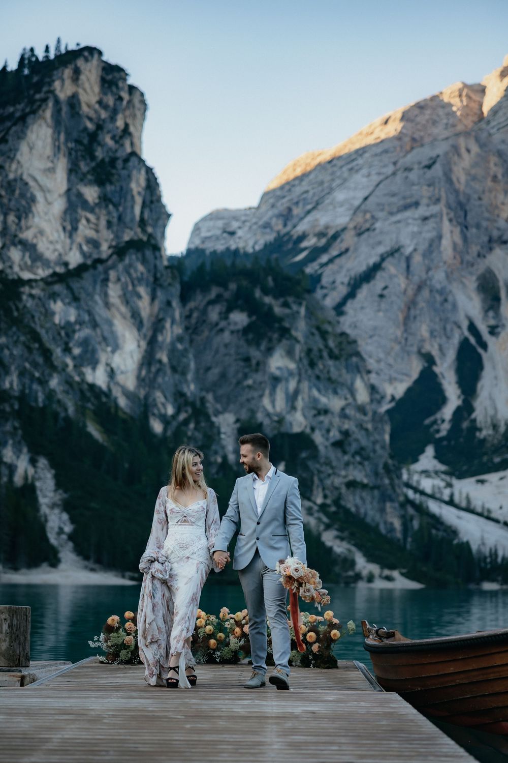 Newlyweds in elegant attire pose on a lakeside dock with dramatic mountain peaks and wooden boats in the background.