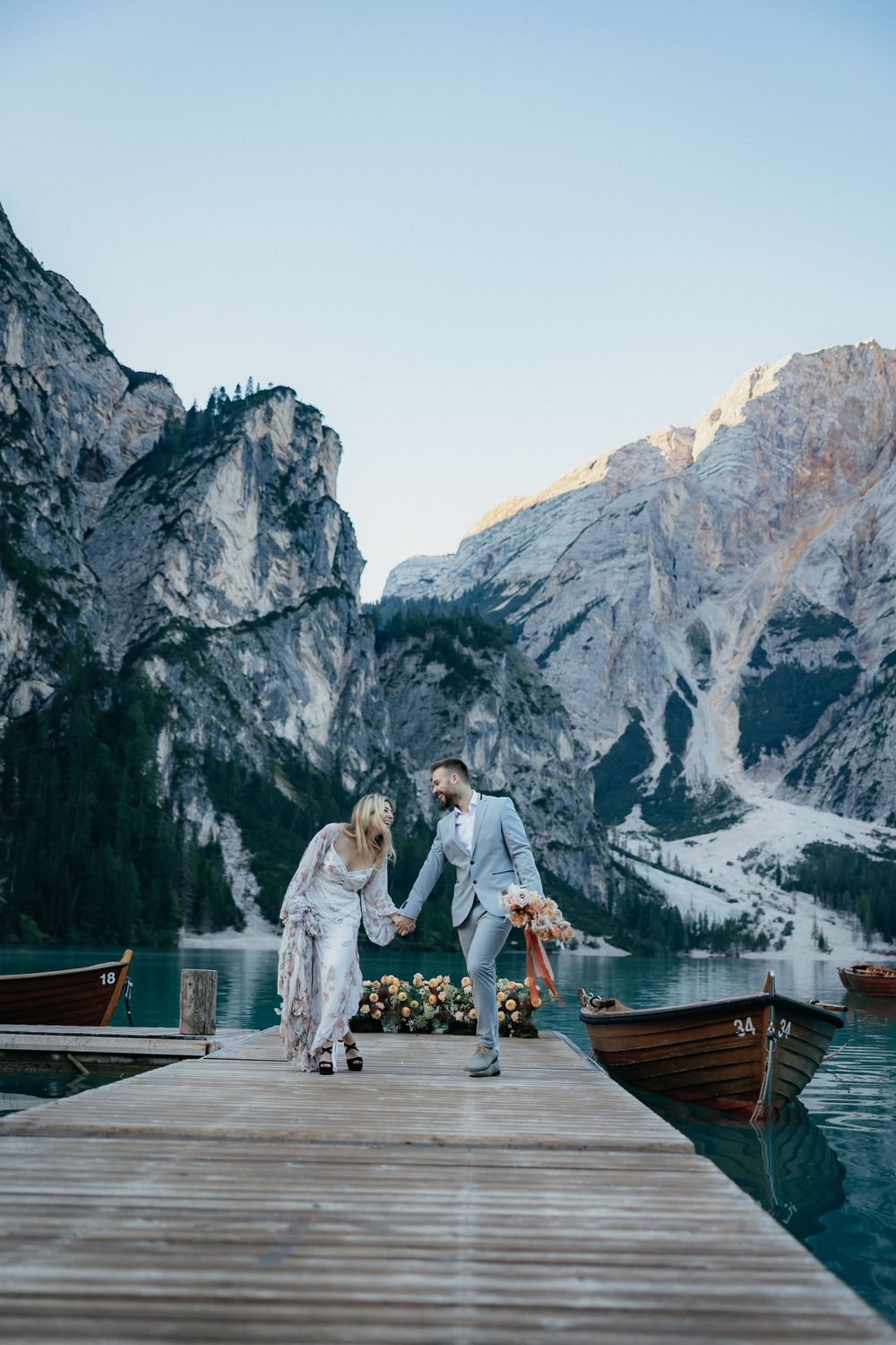 Couple in wedding attire dancing on wooden pier with stunning Dolomites mountains and lake backdrop.
