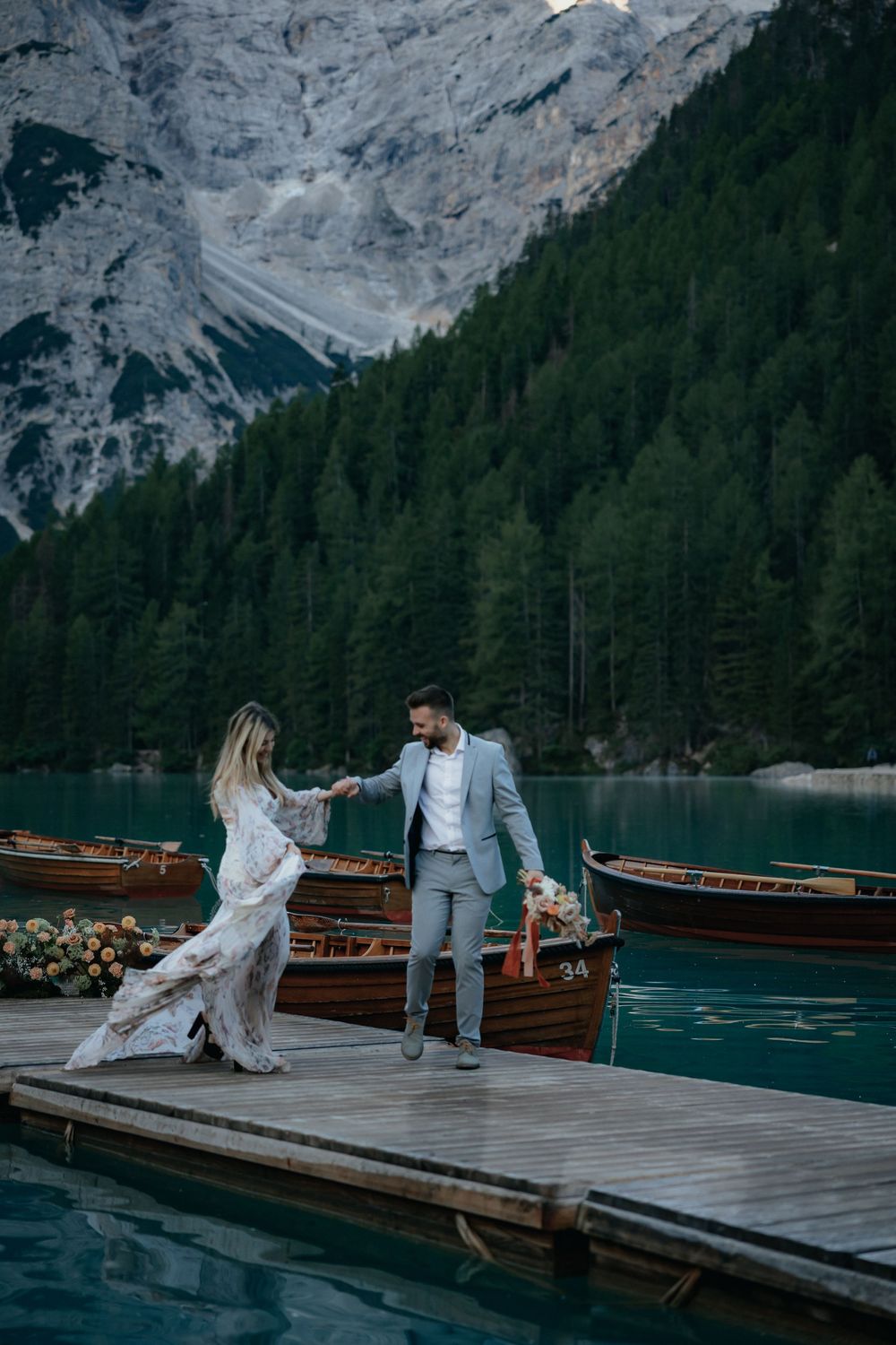 A couple shares a romantic moment on a wooden pier with turquoise alpine lake and towering mountain cliffs behind them.