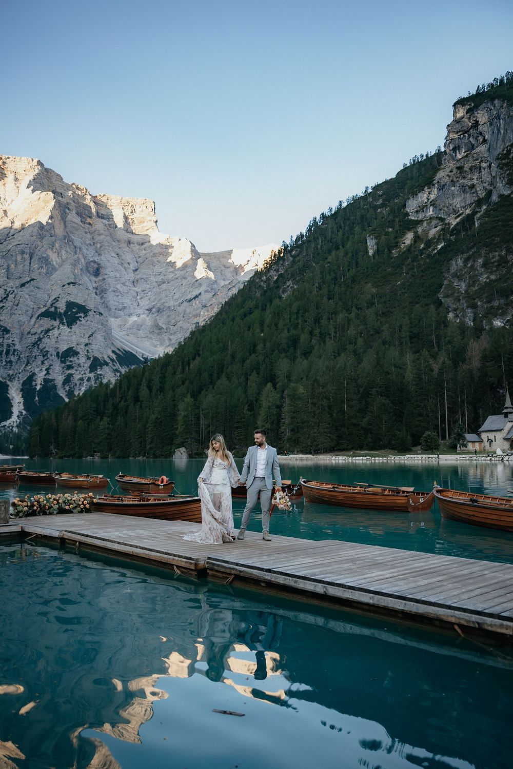 A wedding couple stands on a reflective lake dock surrounded by pine forests and dramatic mountain scenery.