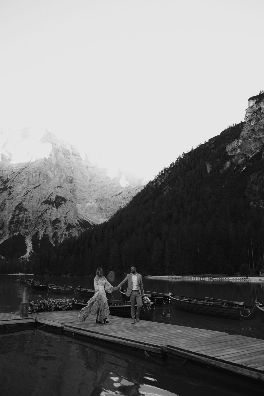 Black and white photograph of wedding scene on mountain lake dock with dramatic alpine scenery.